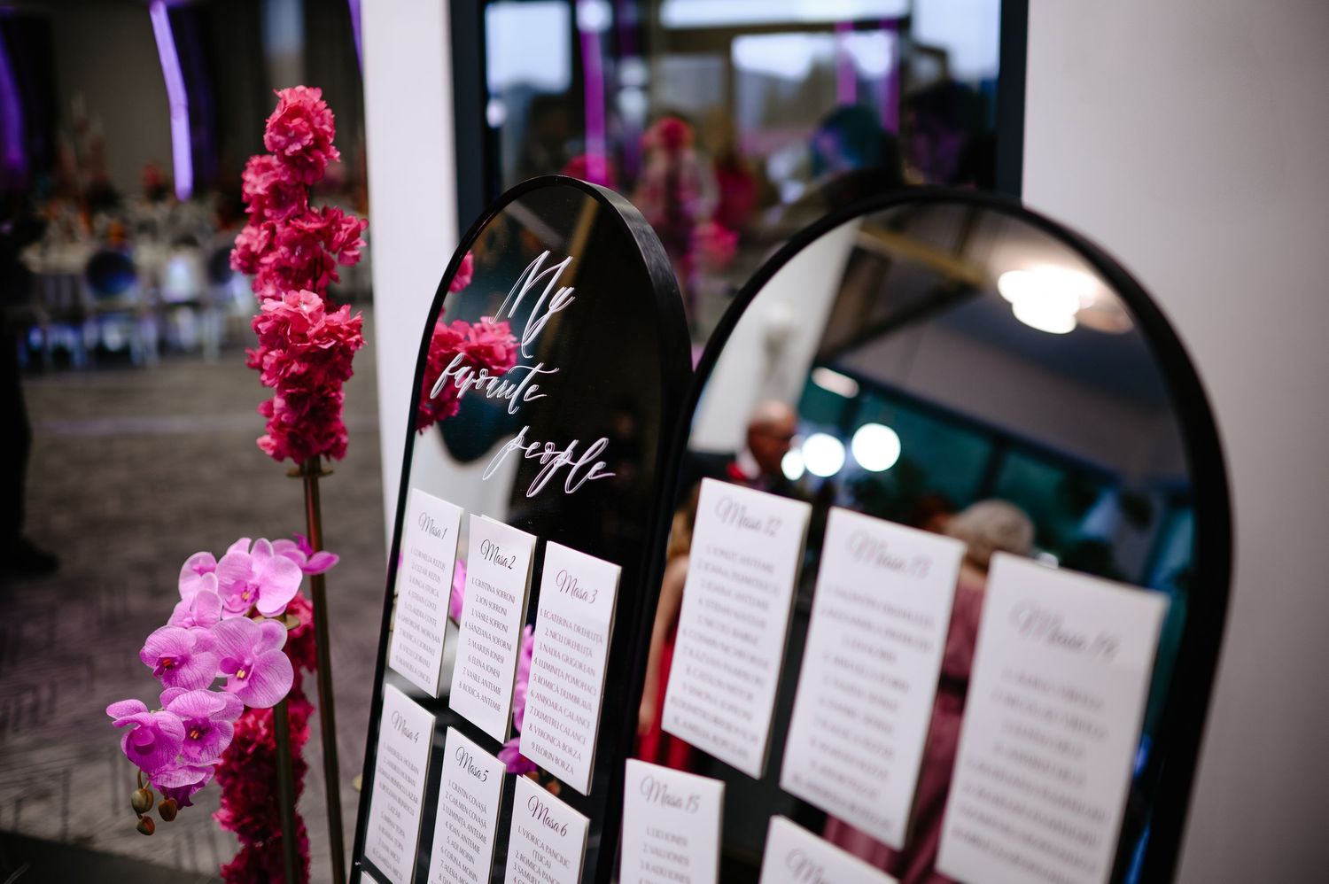 Elegant wedding seating chart display on black mirror with pink orchids and floral arrangements at reception venue.