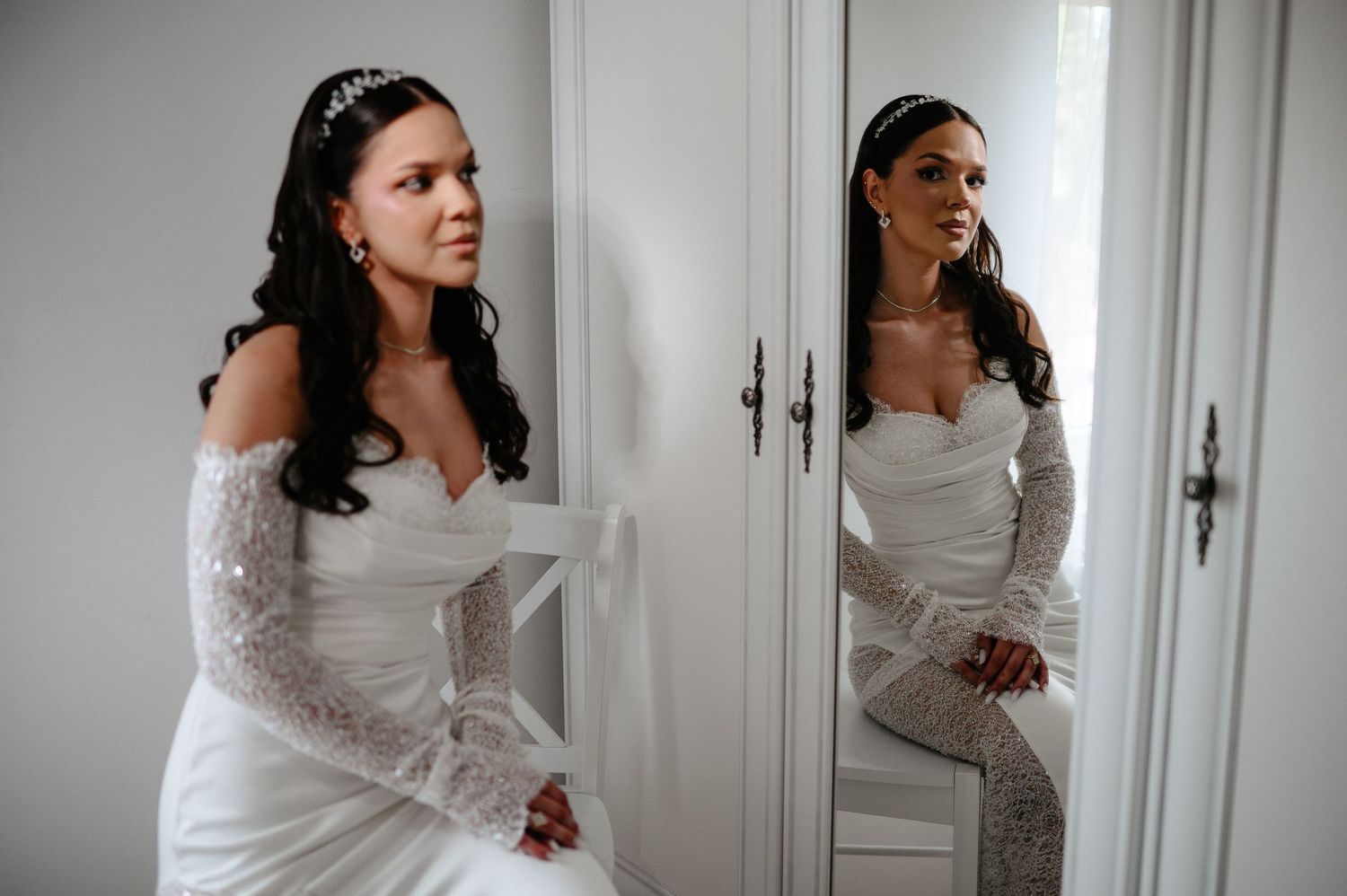Bride in off-shoulder lace wedding dress sitting near wardrobe mirror reflecting her elegant look.
