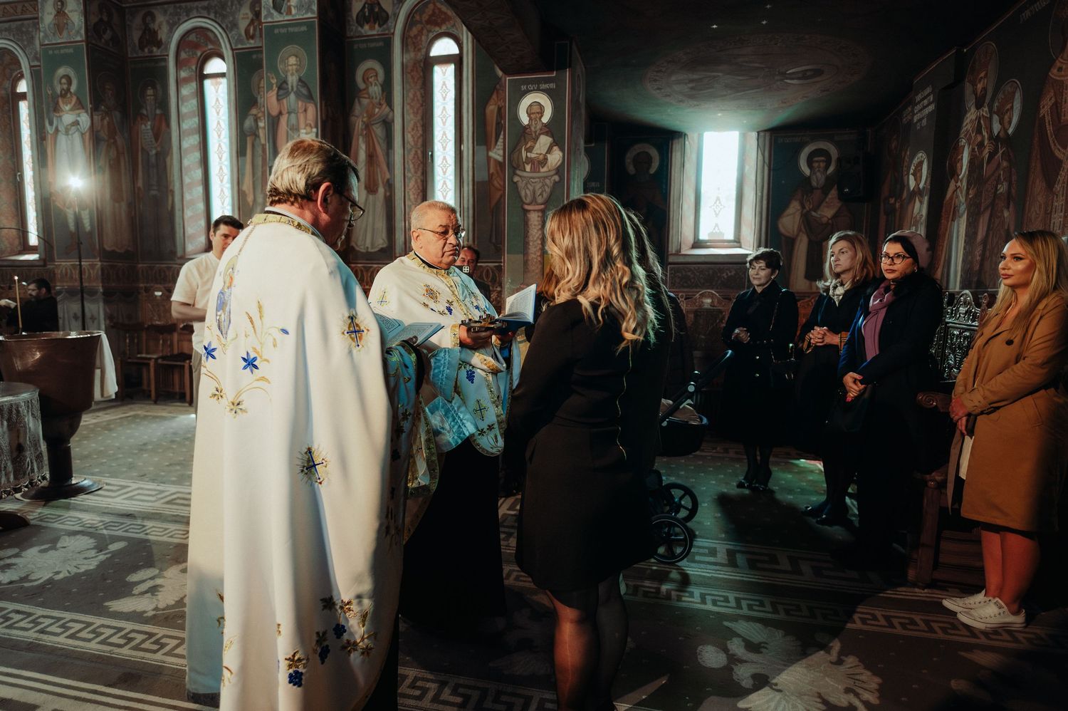 Orthodox priest performing a baptism ceremony with family gathered in a decorated church.