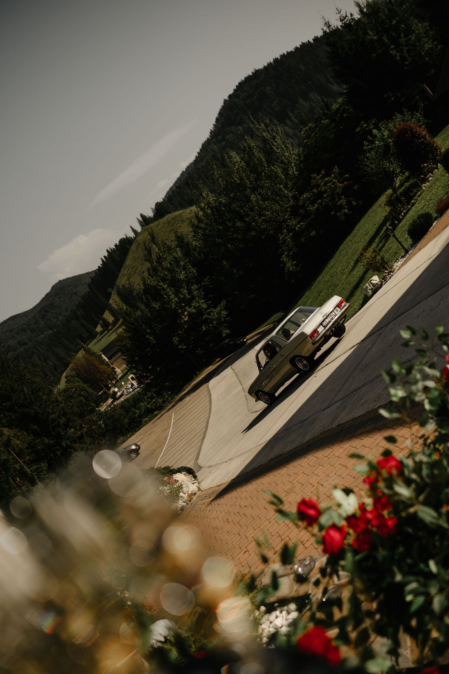 Vintage white car parked on a driveway surrounded by lush green mountains and red roses in the foreground.