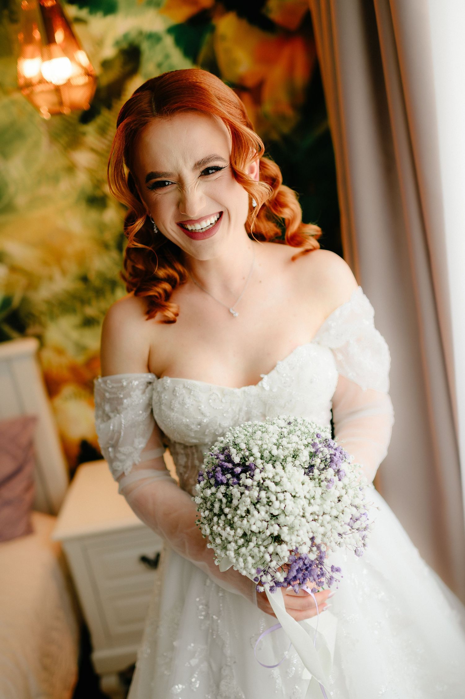 Smiling redhead bride in off-shoulder lace gown holding white and purple baby's breath bouquet indoors.