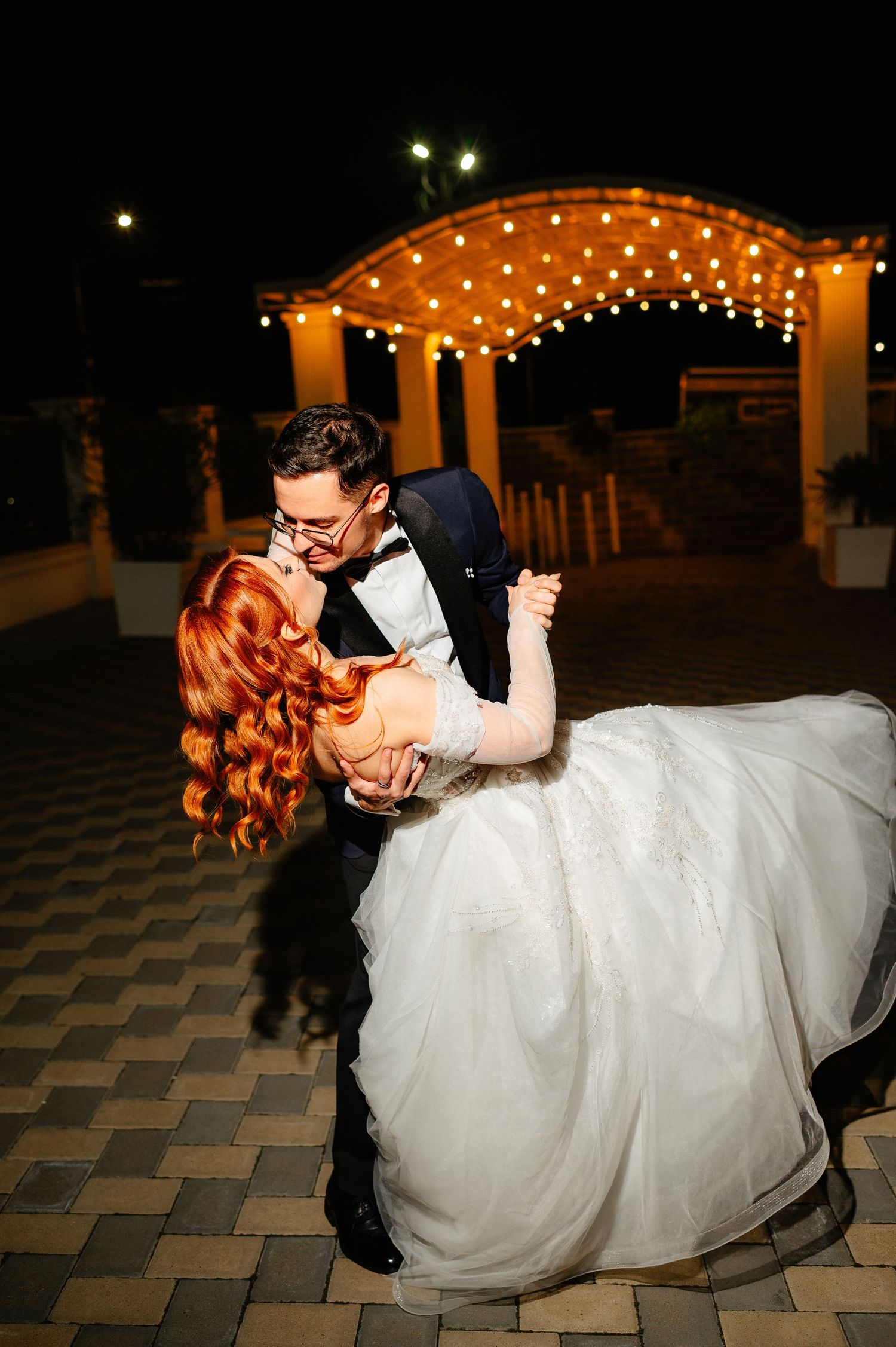 Groom dips bride in white gown under string lights at night during romantic wedding first dance.