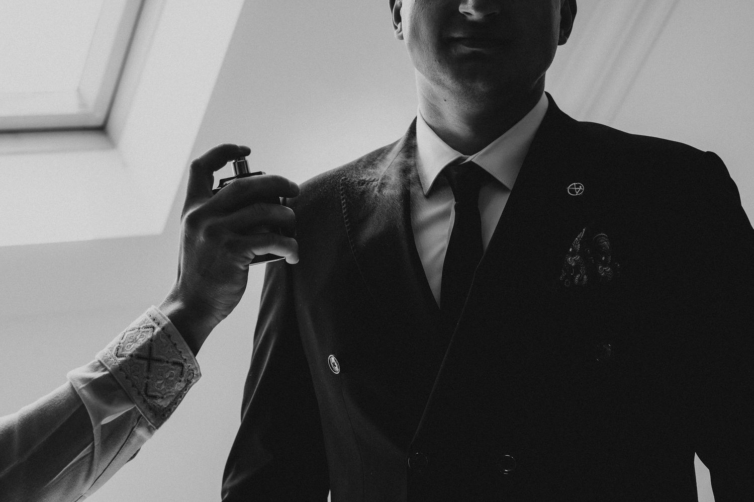 Black and white photo of a man in a suit being sprayed with cologne by a tattooed hand.