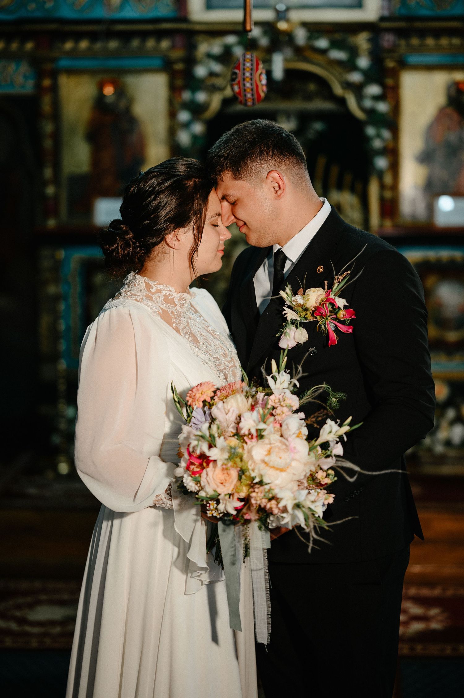Bride and groom touch foreheads romantically inside ornate Orthodox church, bride holding pastel floral bouquet.