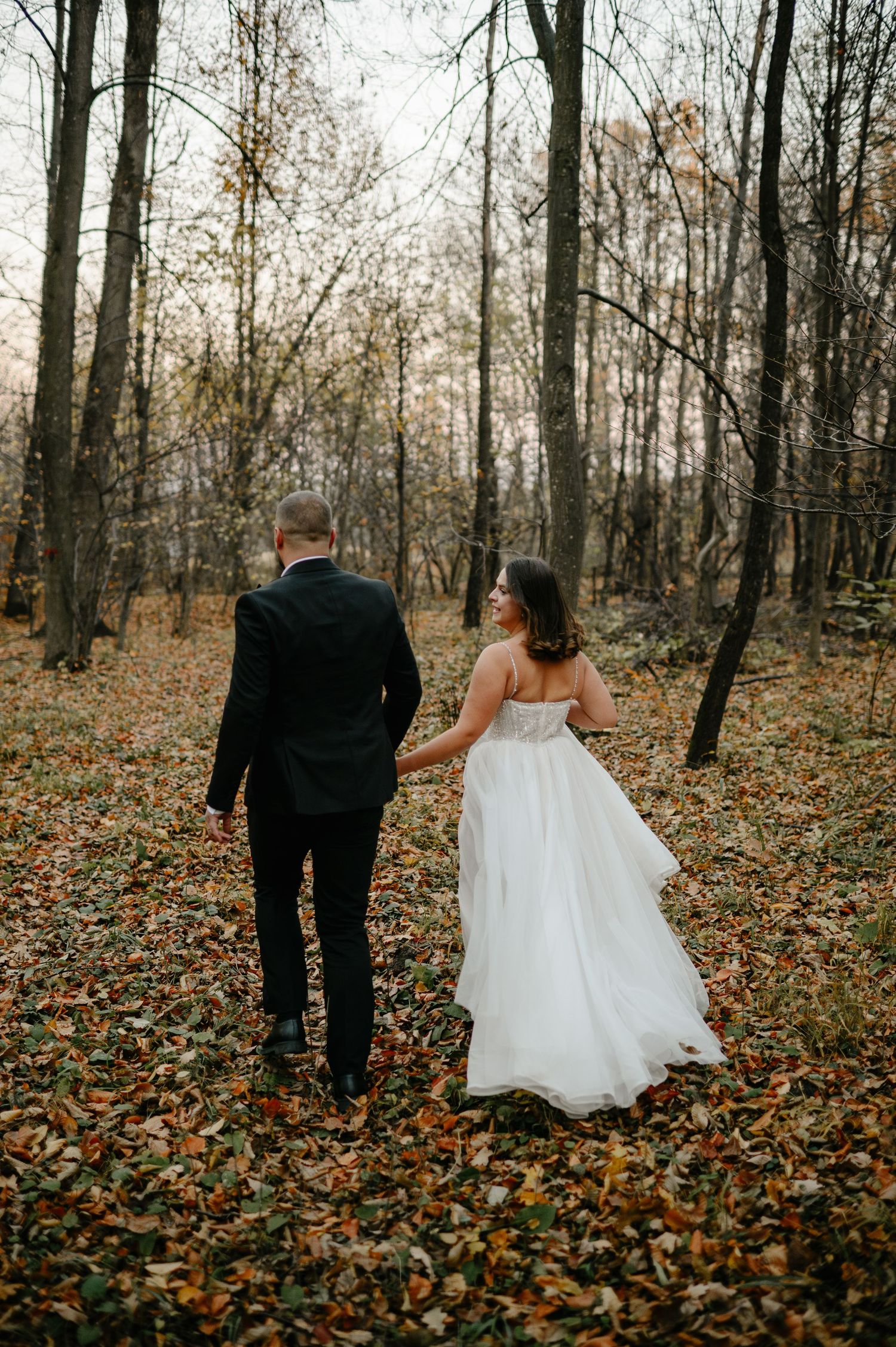 A bride and groom walk hand in hand through an autumn forest, surrounded by fallen leaves and bare trees.