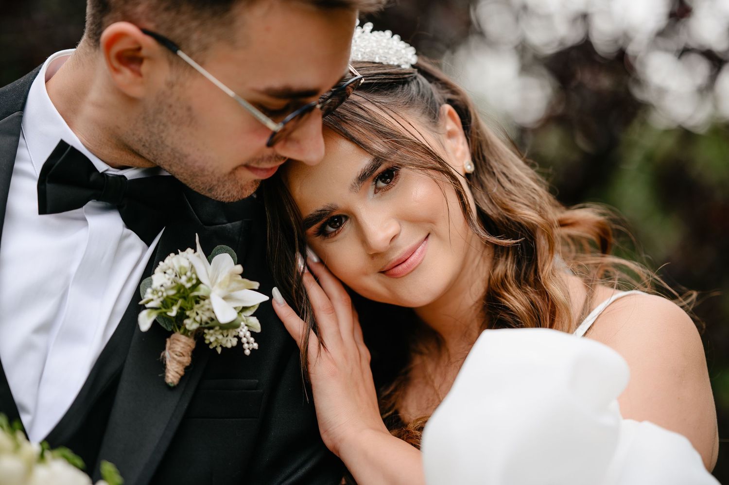 Romantic wedding couple portrait with groom in black tuxedo kissing bride's forehead, bride smiling in white gown.