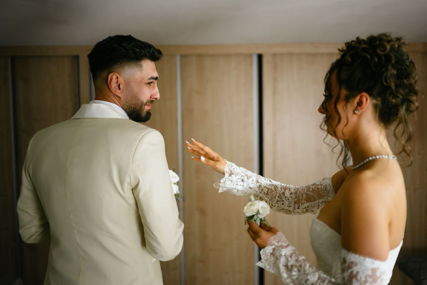 Bride in lace gown touching groom's face in cream suit during wedding preparation indoors.