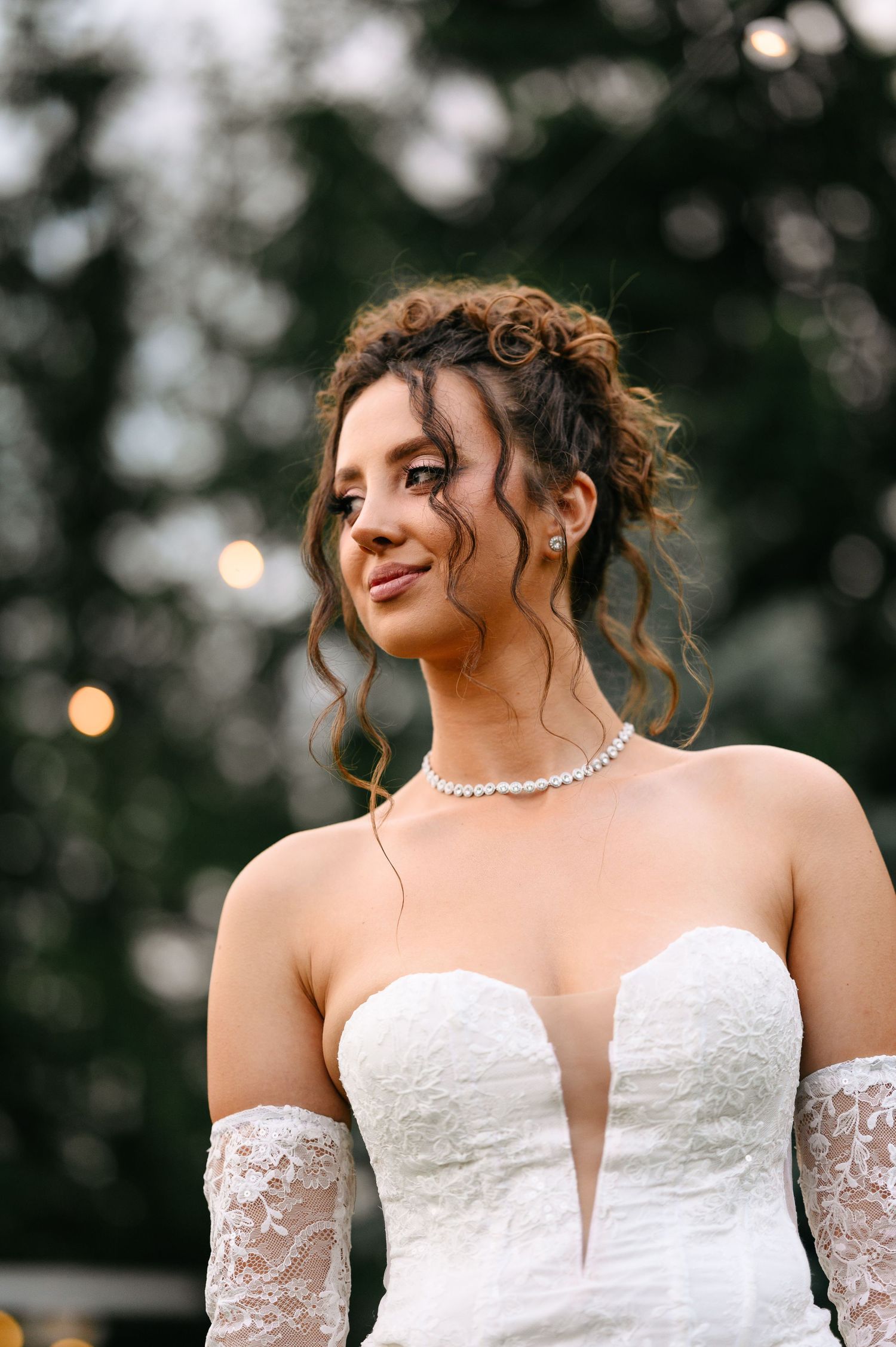 Bride in strapless lace wedding gown with diamond necklace and lace gloves, curly updo hairstyle, outdoor wedding.