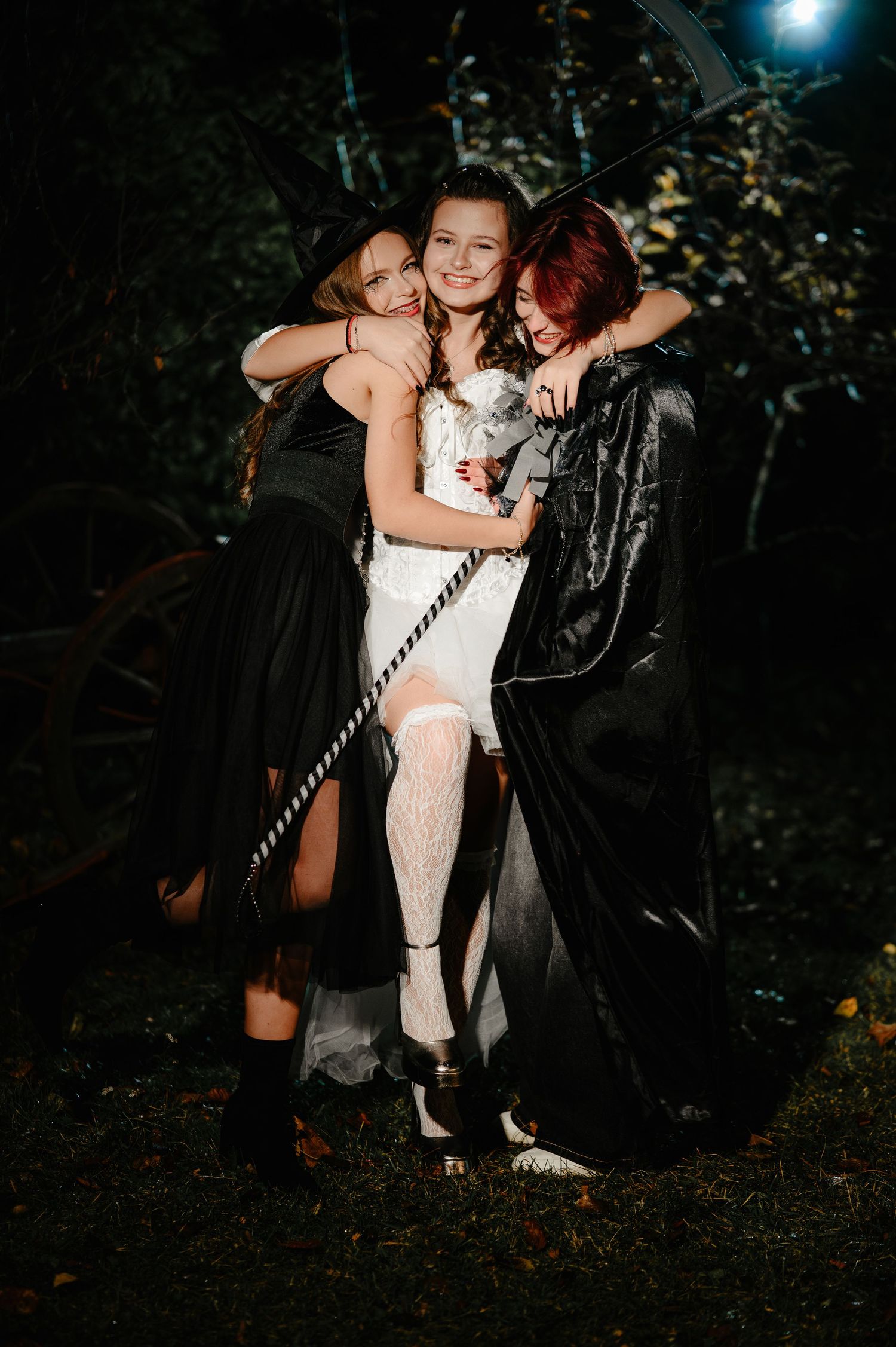 Three young women in Halloween costumes hugging outdoors at night, two in black capes and one in white.