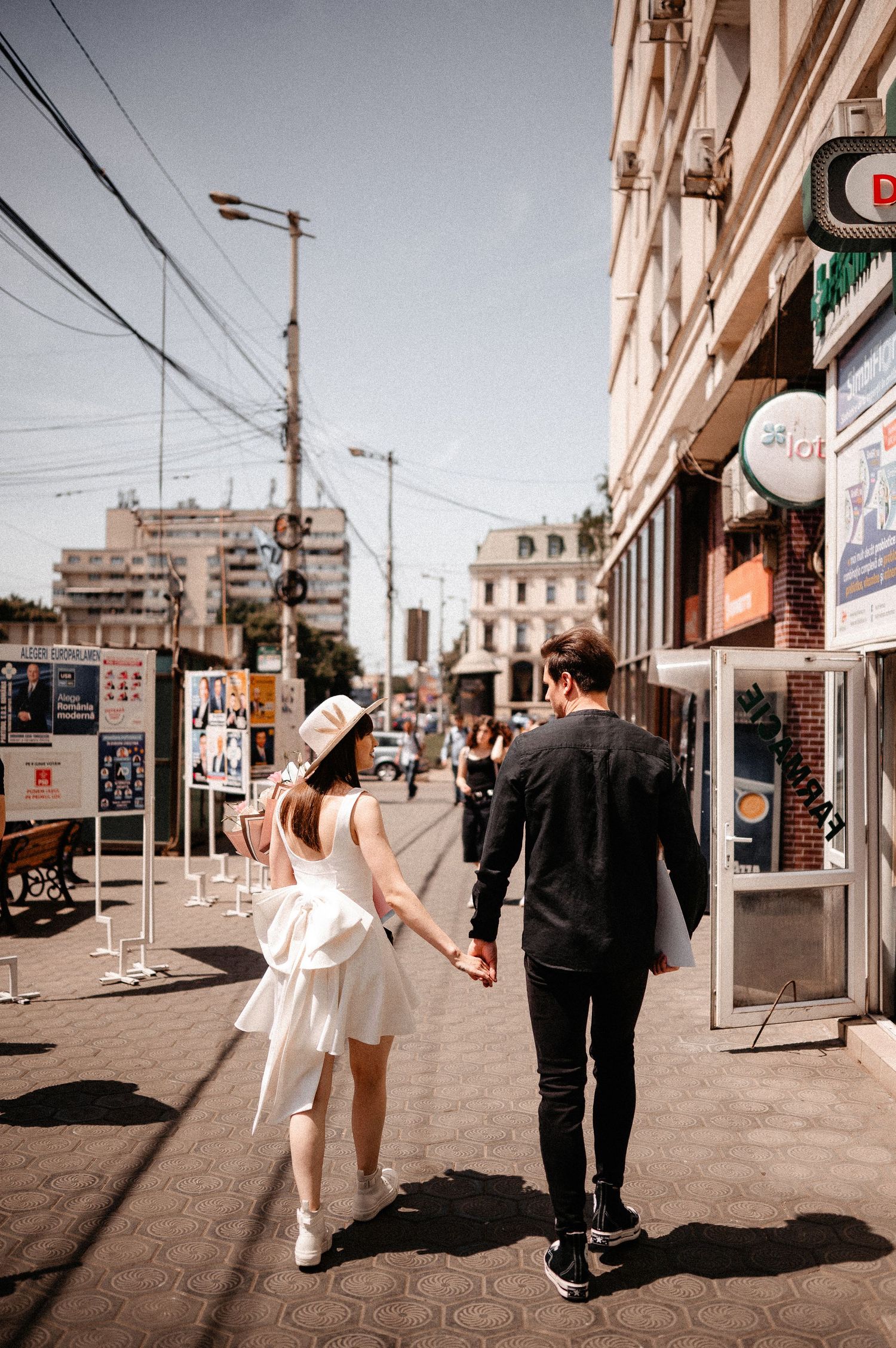 Young couple holding hands walking down a sunny urban street, woman in white dress and hat, man in black outfit.