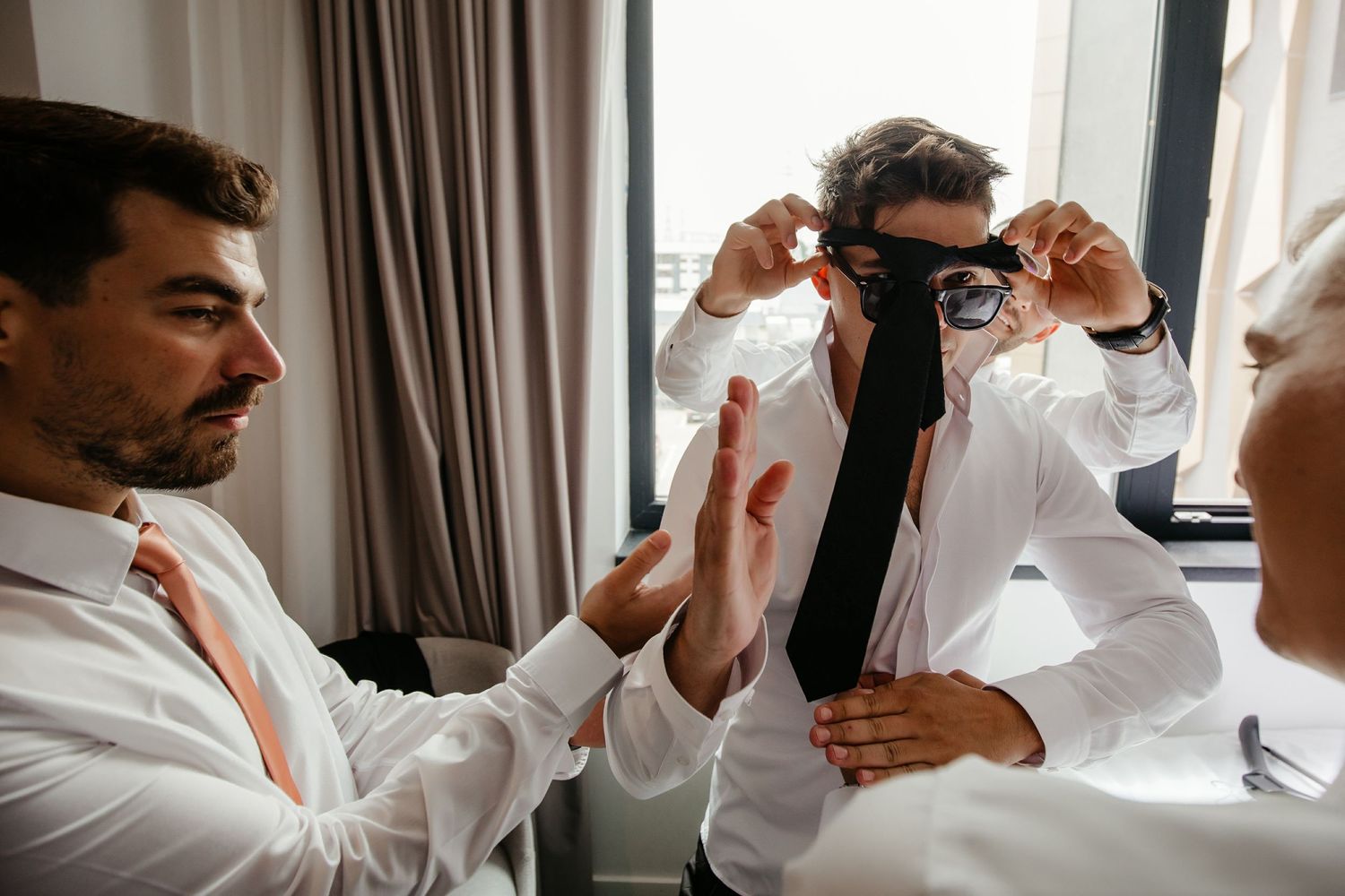 Groomsmen help groom adjust black tie while getting dressed for wedding day preparations.