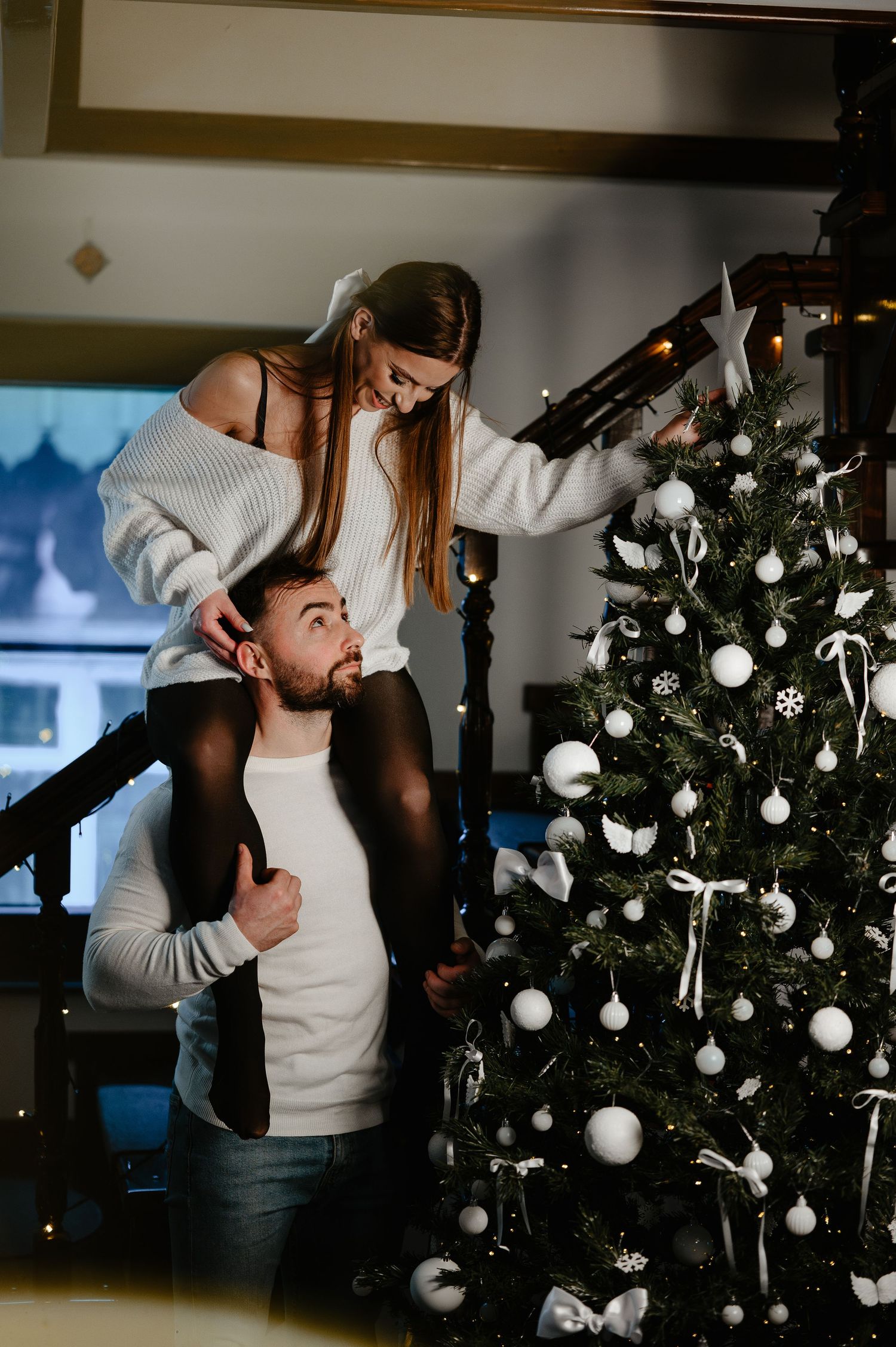 Couple decorating a white-ornament Christmas tree together, woman perched on man's shoulders smiling.