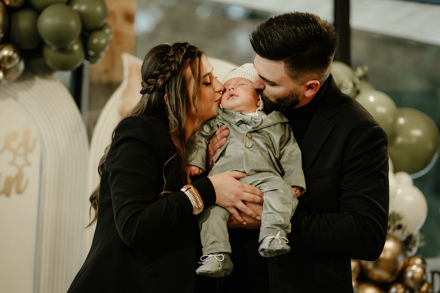 Loving parents kissing newborn baby together at colorful balloon-decorated baby shower celebration.