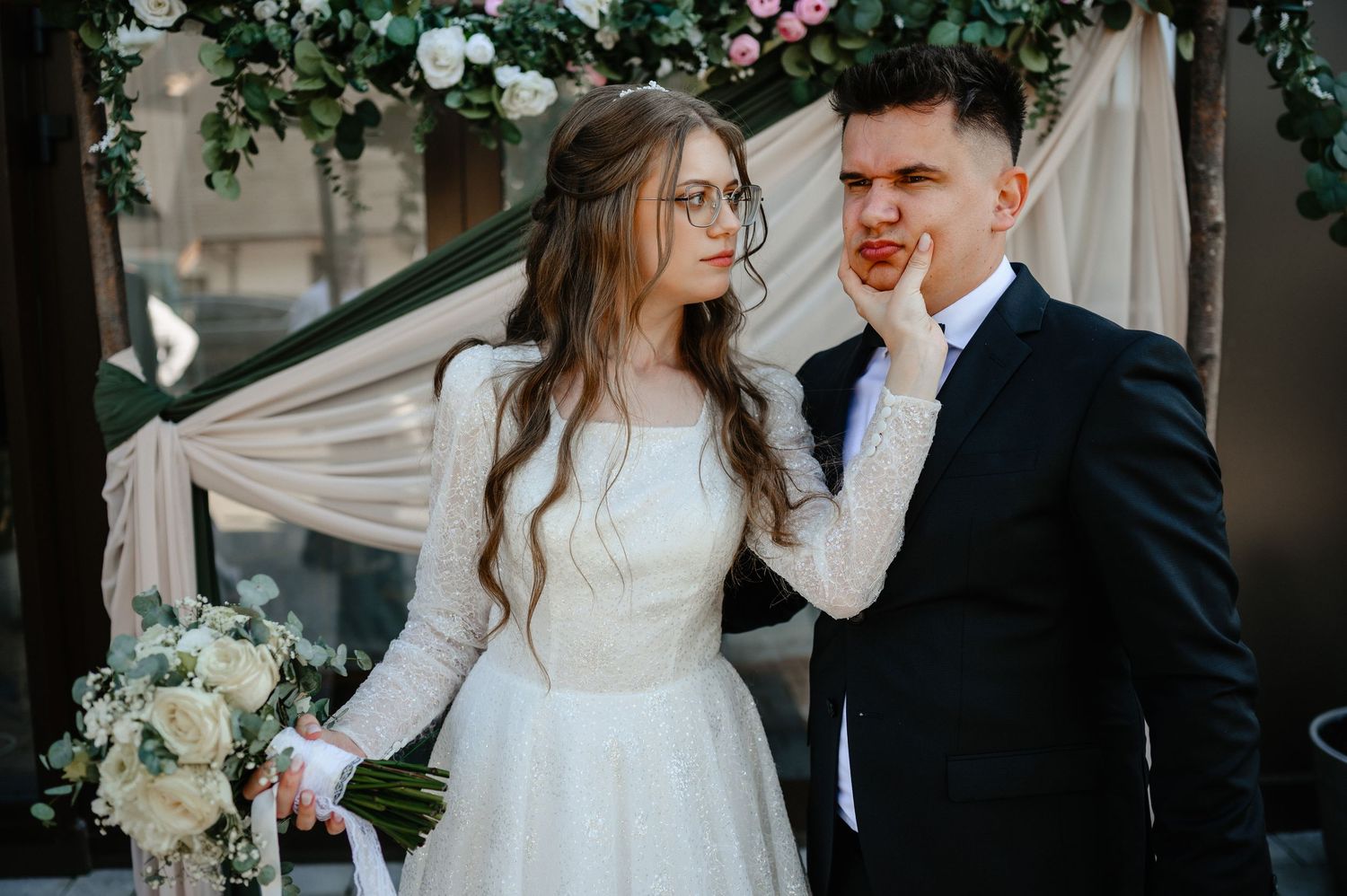 Bride in white lace dress gently touching grooms face while holding a bouquet outdoors.