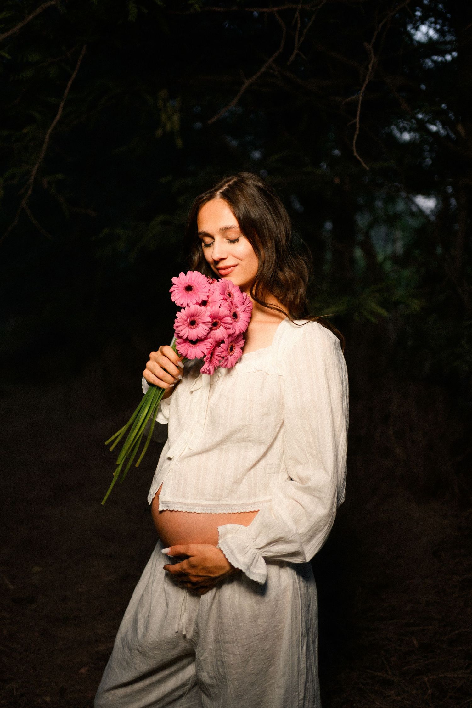 Pregnant woman in white outfit holding pink gerbera daisy bouquet, smiling with eyes closed in a sunlit forest setting.