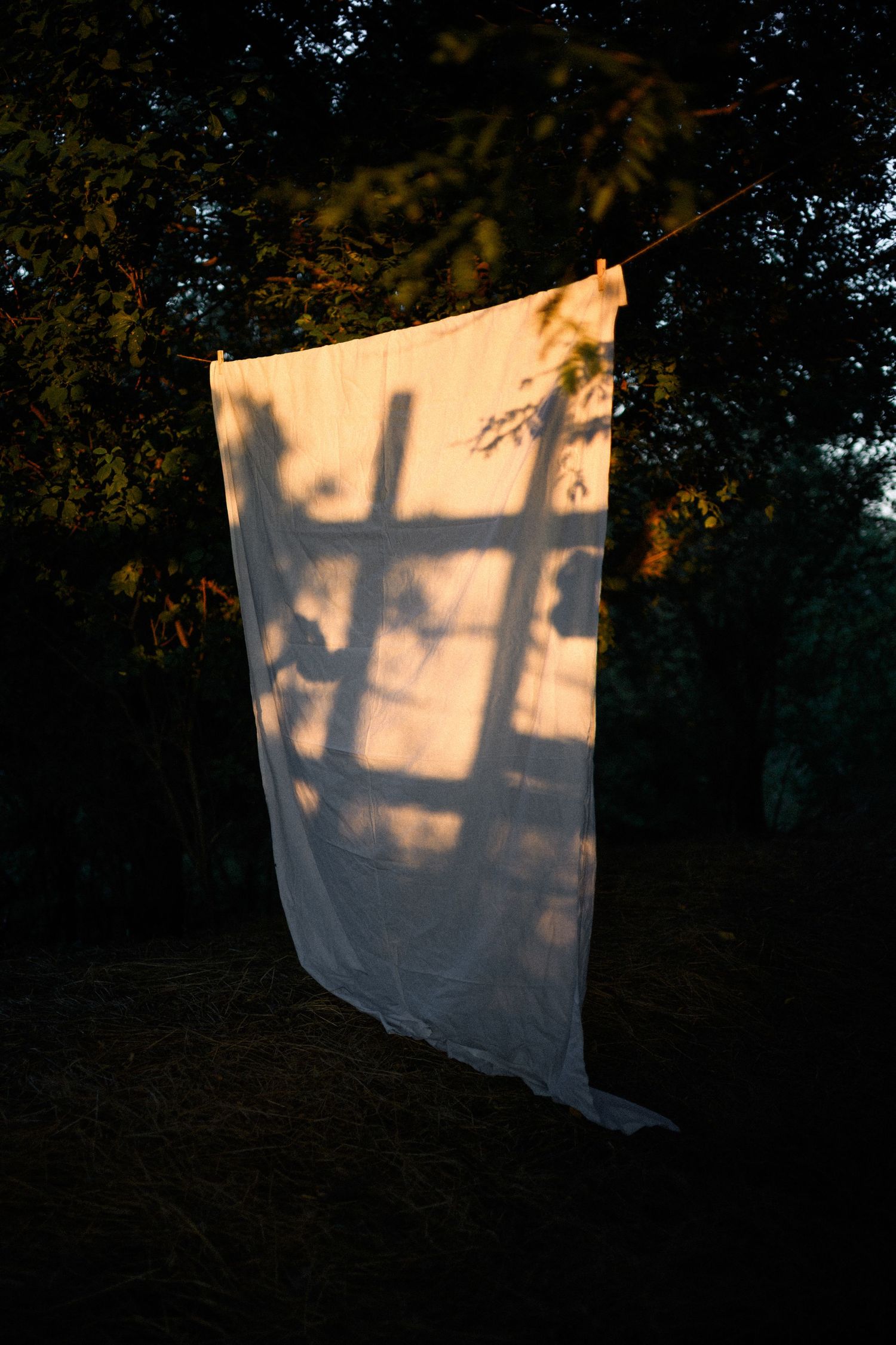 White sheet hanging on clothesline at dusk with shadow of wooden ladder cast by golden sunset light.