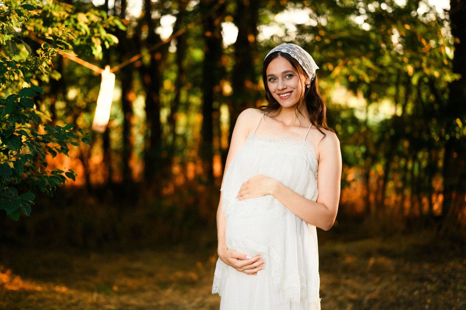 Smiling pregnant woman in white dress cradling baby bump in sunlit forest during golden hour maternity photoshoot.