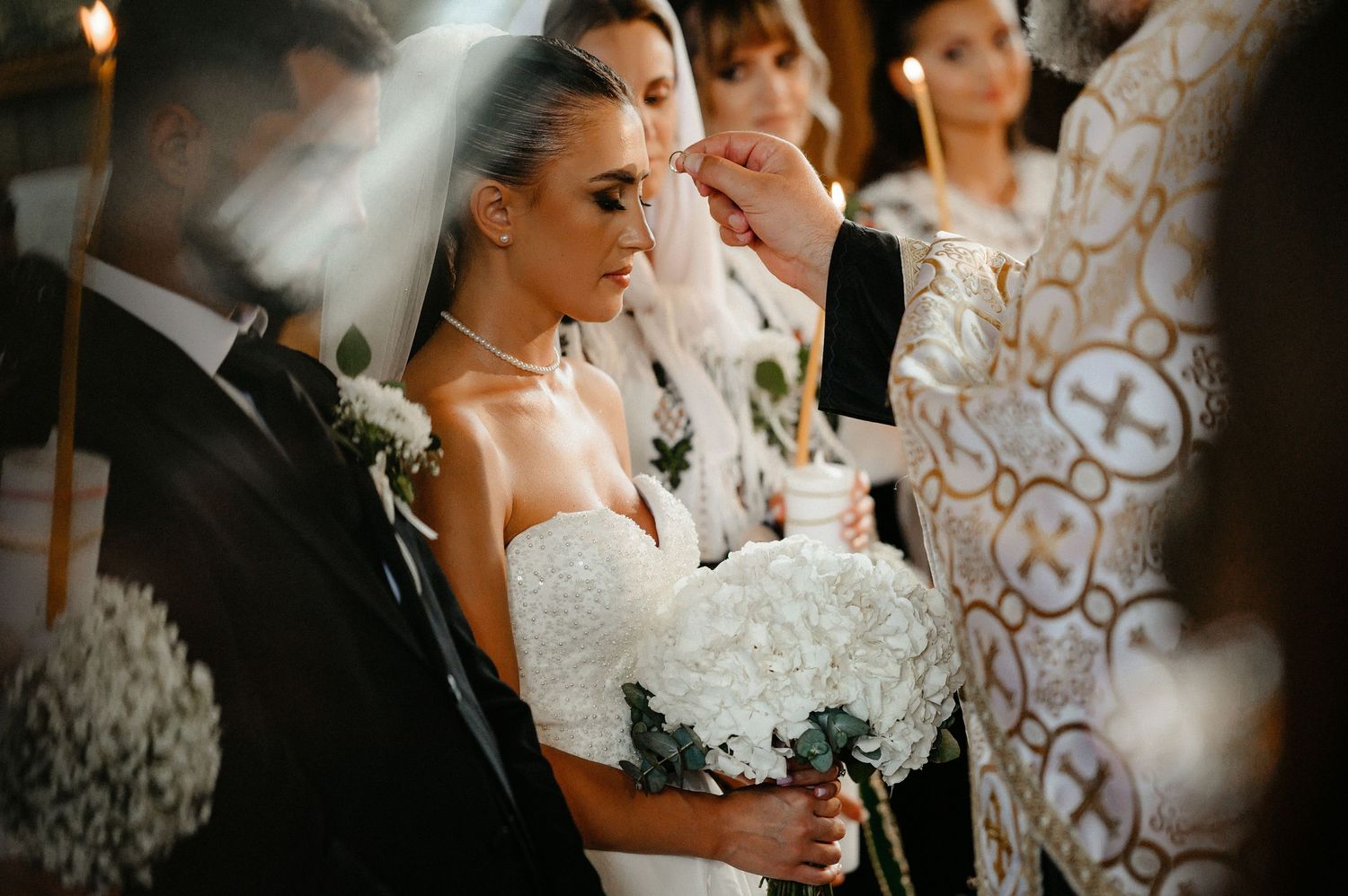 Bride in white lace gown holding hydrangea bouquet receives blessing from Orthodox priest during wedding ceremony.