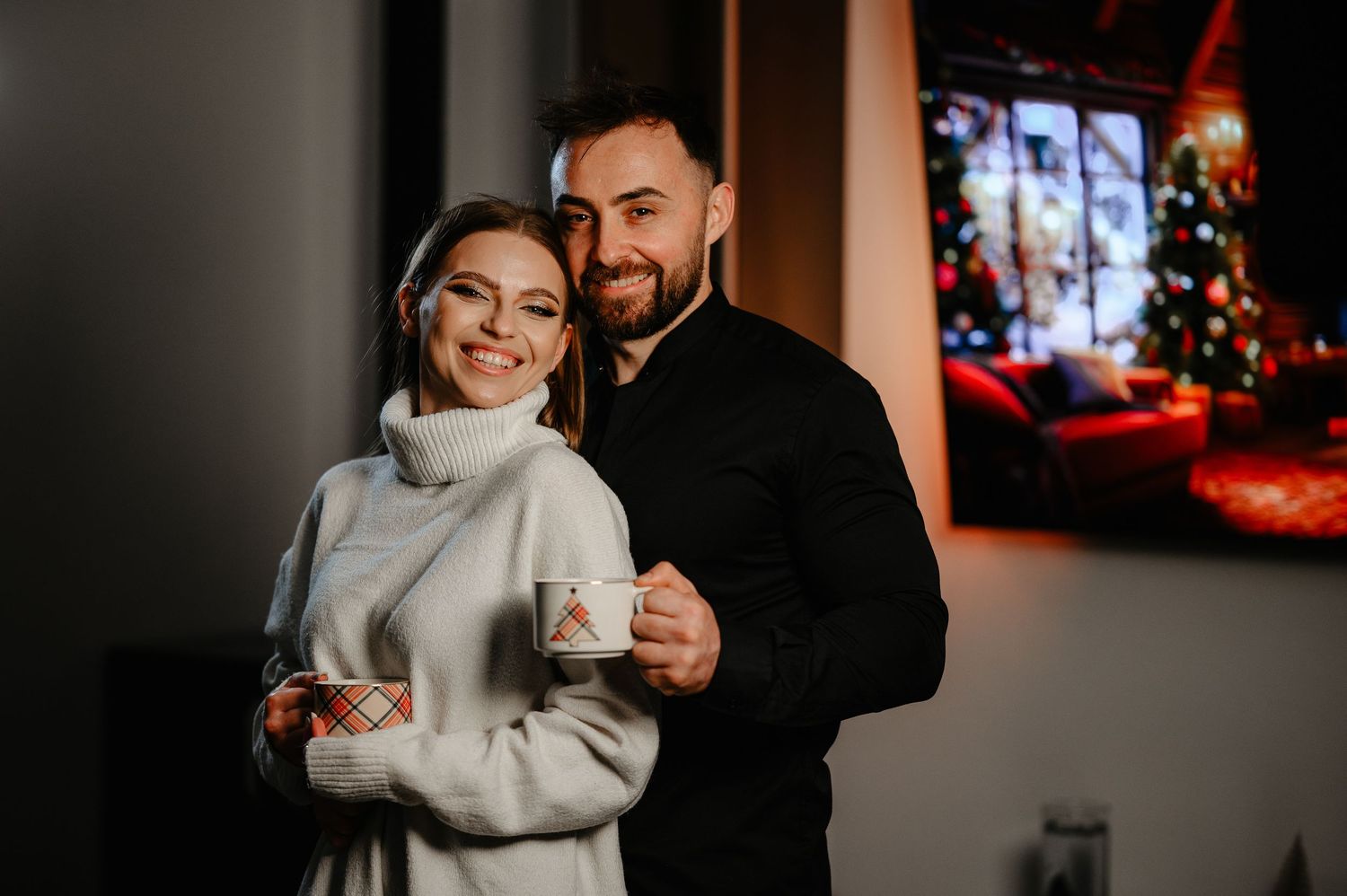 Happy couple holding festive mugs in front of a Christmas tree display at home.