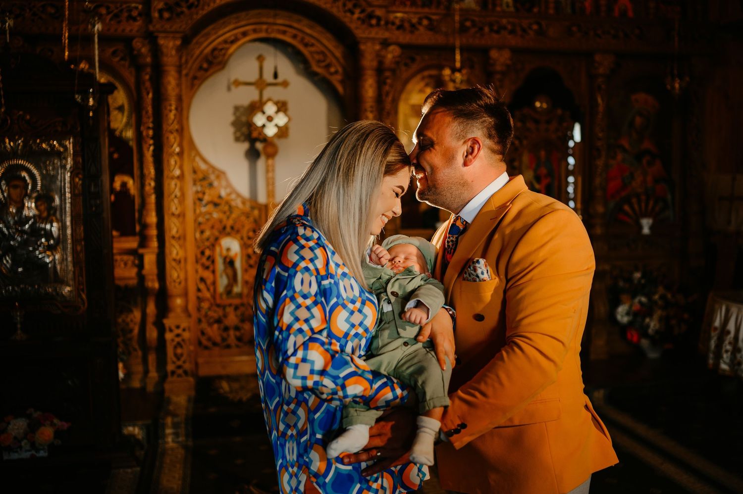 Parents sharing tender moment with newborn baby in front of golden Orthodox church altar.