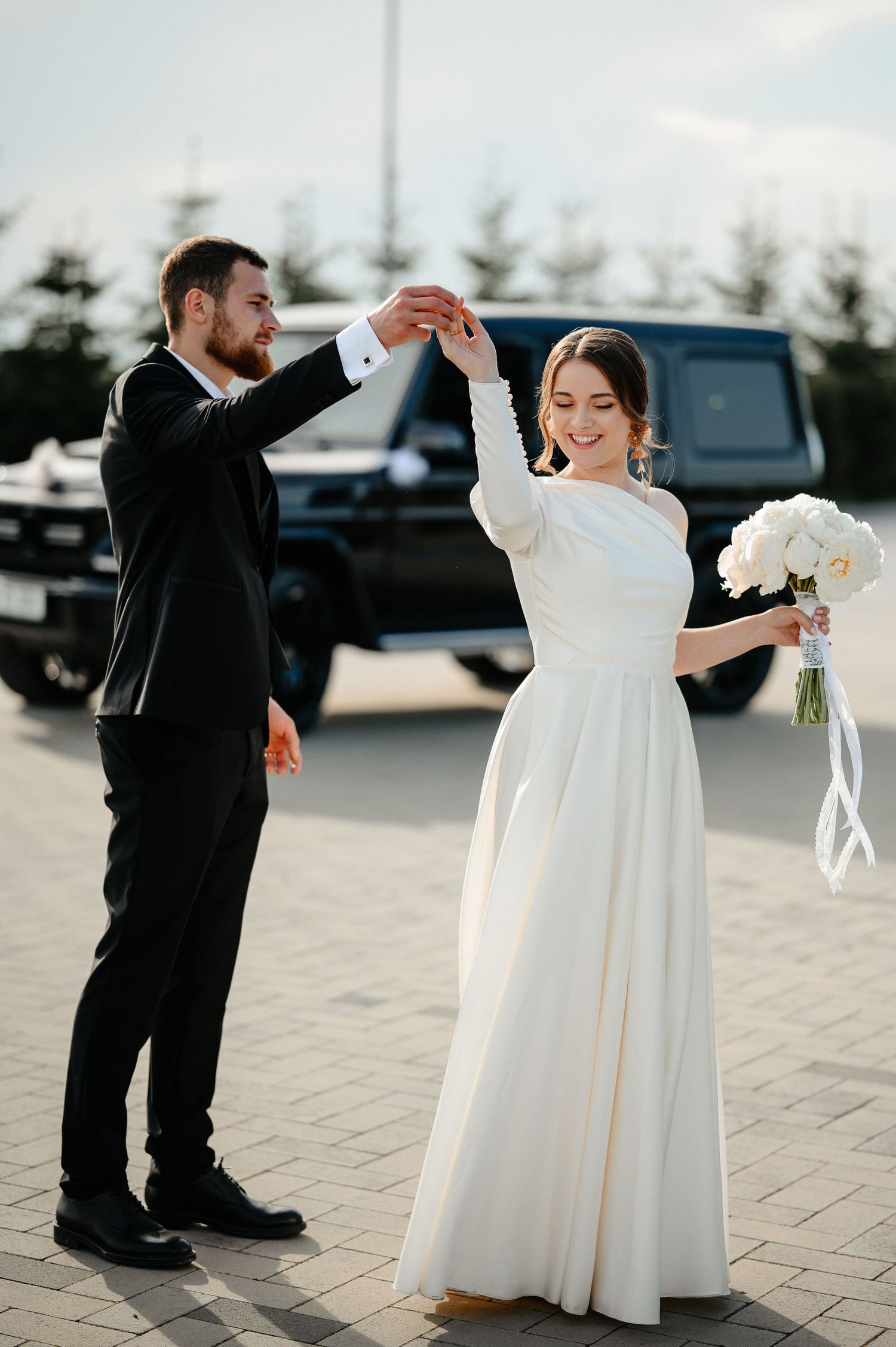 Bride and groom dancing outdoors near black SUV, bride in white gown holding bouquet.