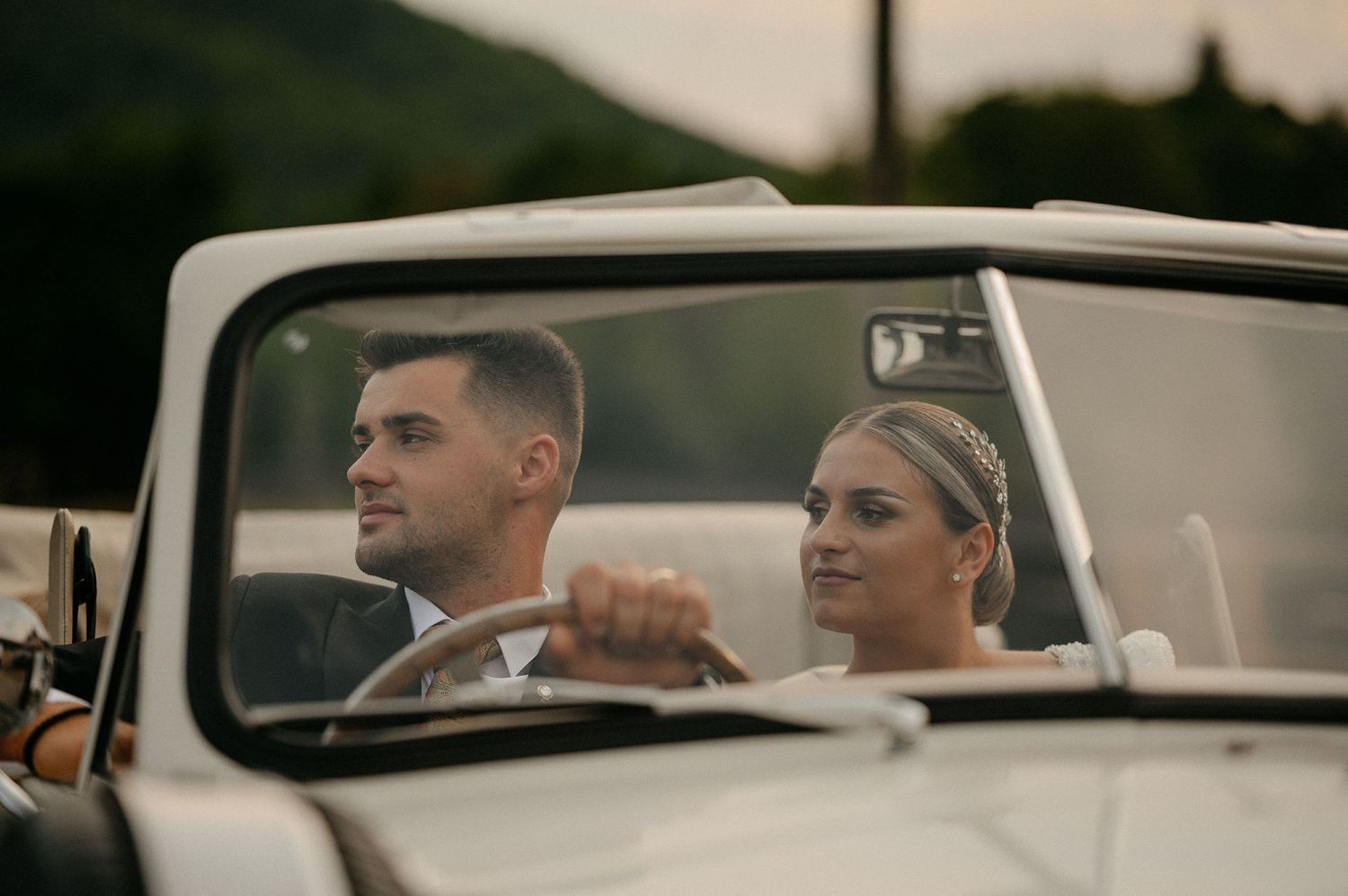 Newlywed couple riding in a vintage white convertible car through a scenic countryside landscape.