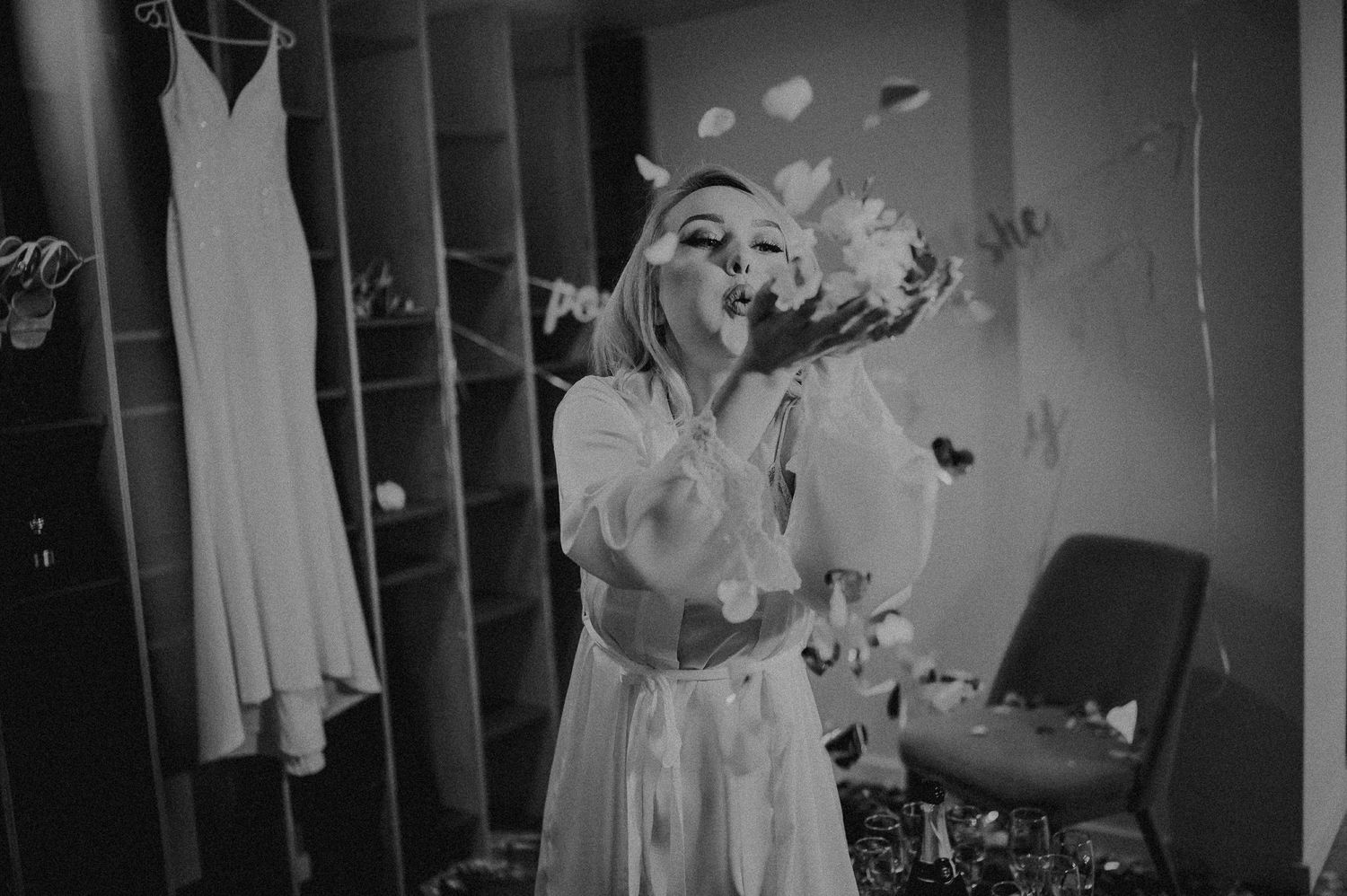 Black and white photo of a bride in a white robe blowing flower petals with her wedding dress hanging in background.