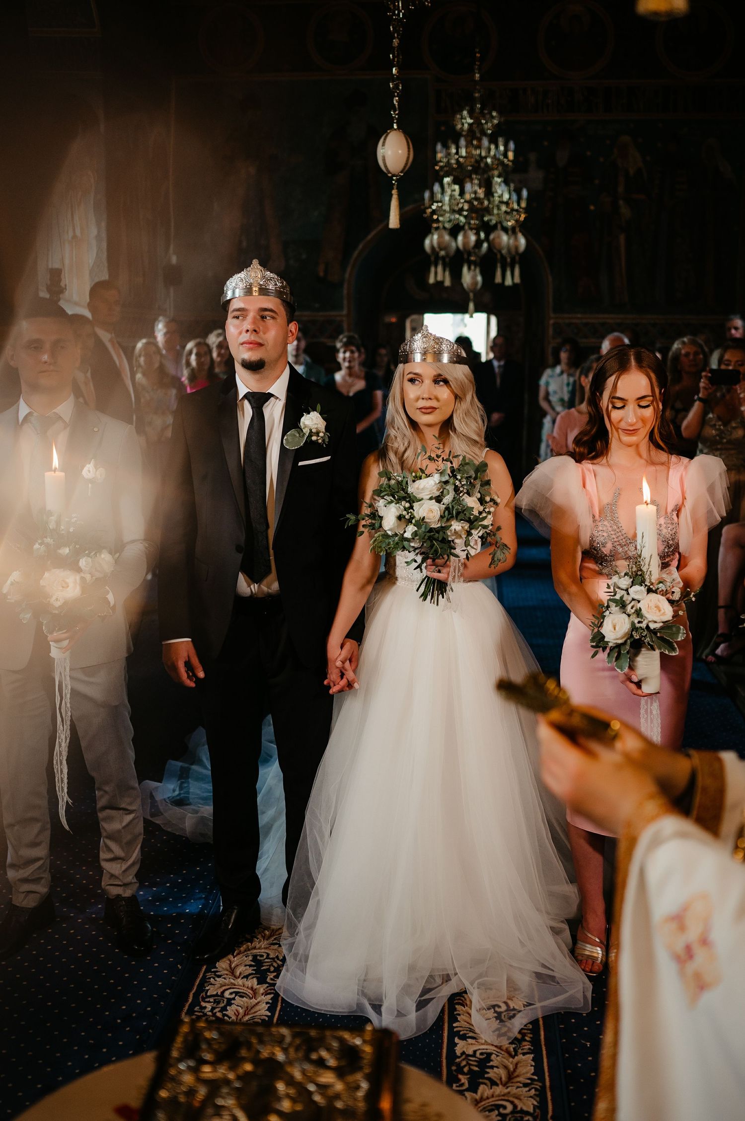 Bride and groom wearing ceremonial crowns holding hands during Orthodox wedding ceremony in ornate church.