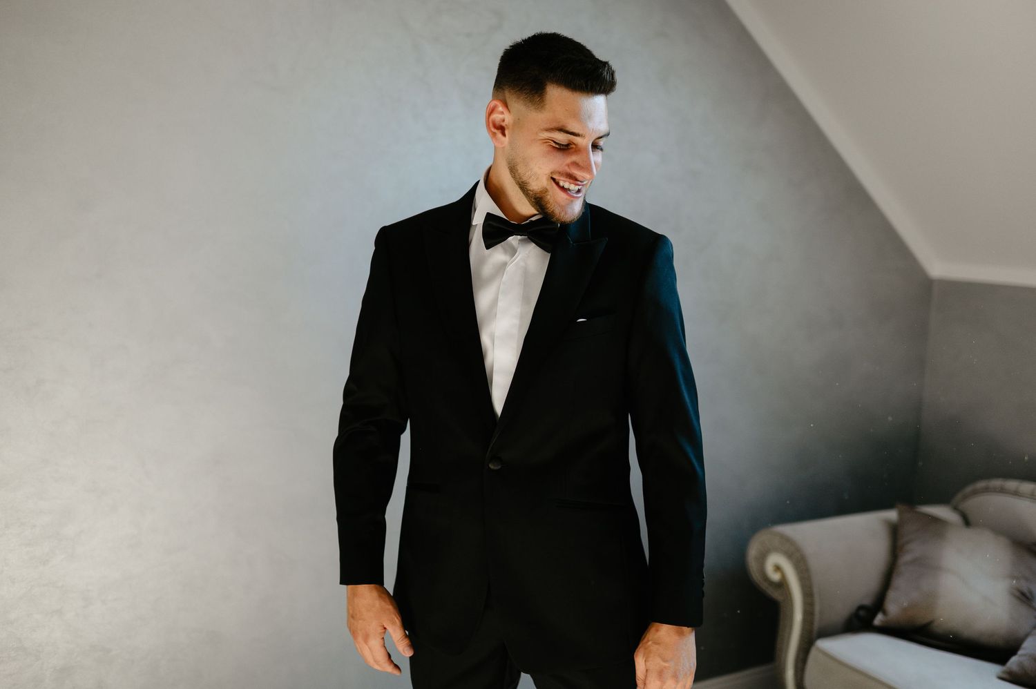 Groom smiling in black tuxedo with bow tie standing in bright room.