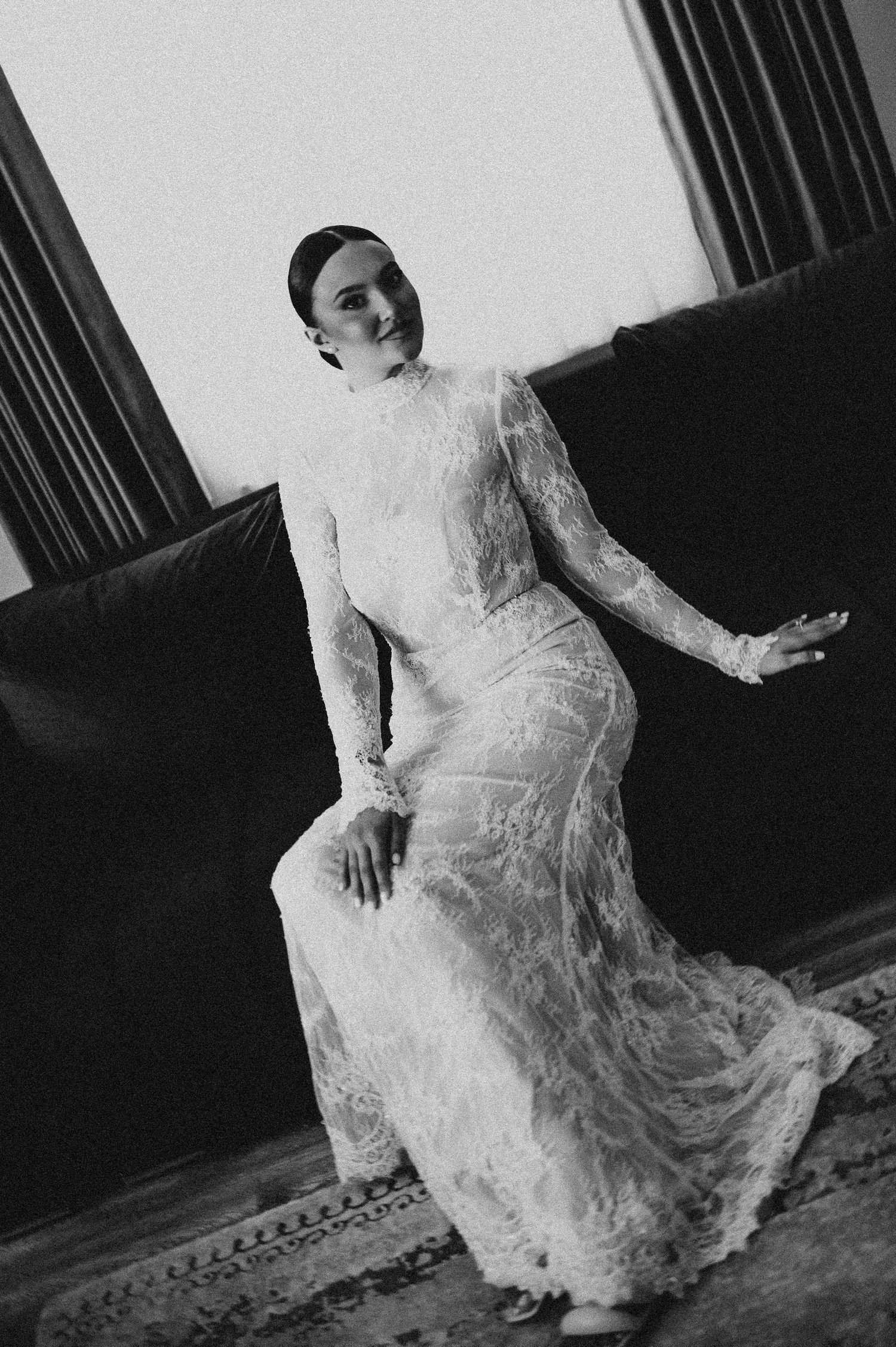 Bride in a long-sleeve lace wedding dress seated on a dark sofa in a dramatic black and white photo.
