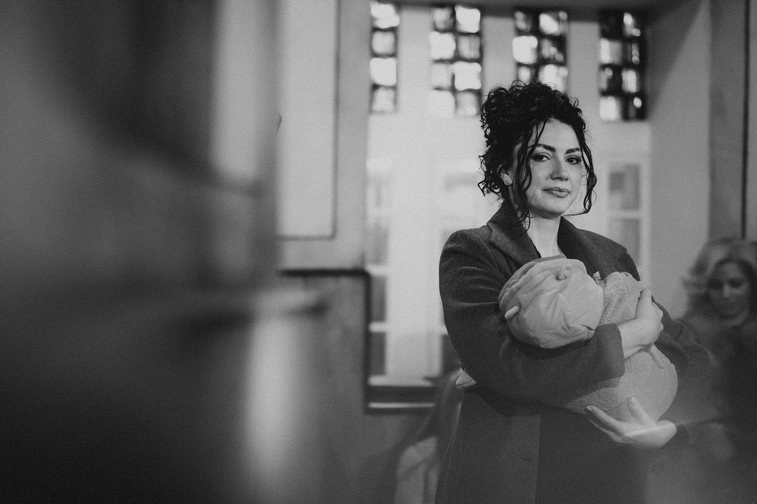 Black and white image of curly-haired woman holding newborn baby inside Orthodox church.
