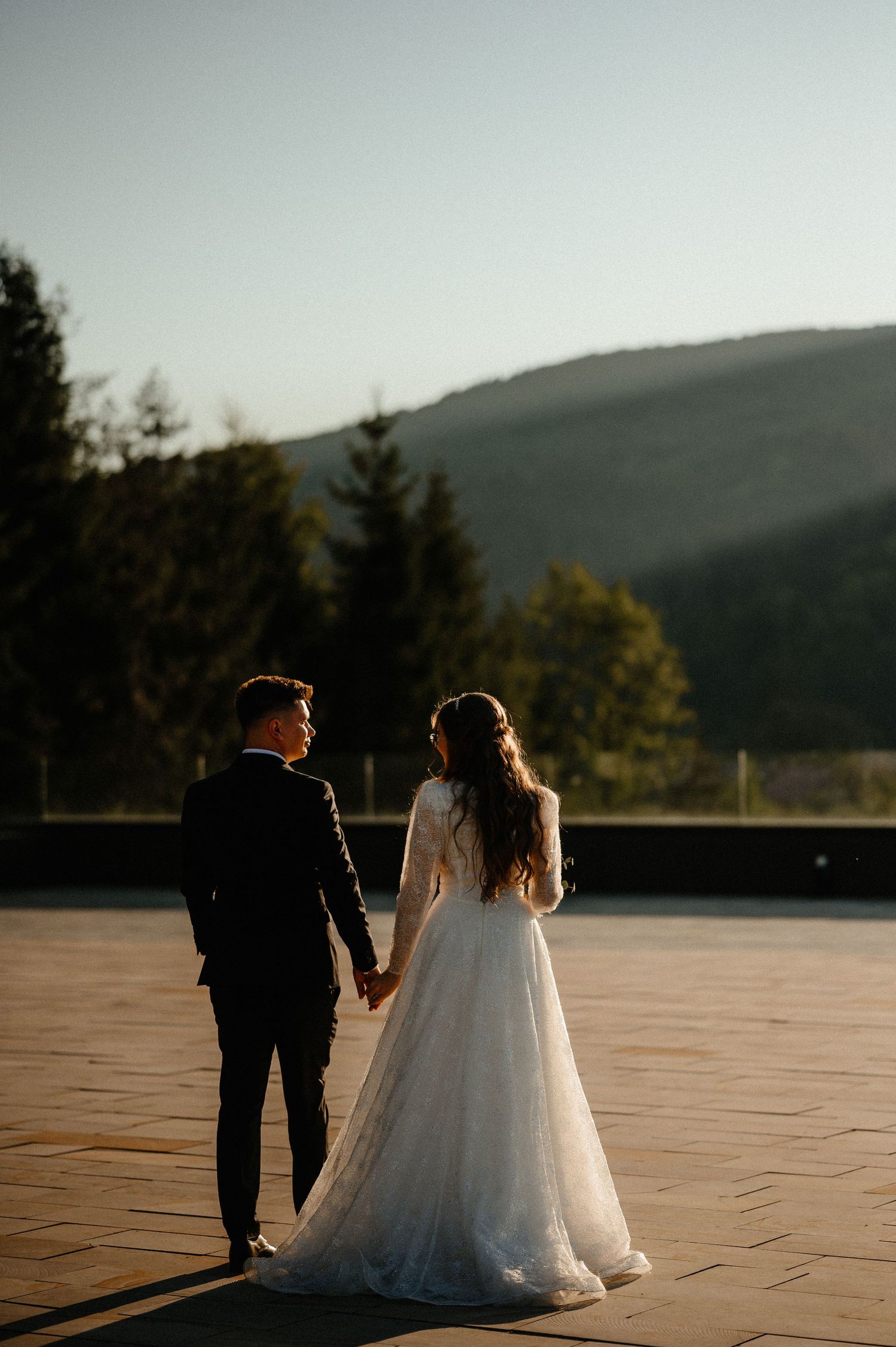 Bride and groom holding hands facing mountain landscape in warm golden evening sunlight.
