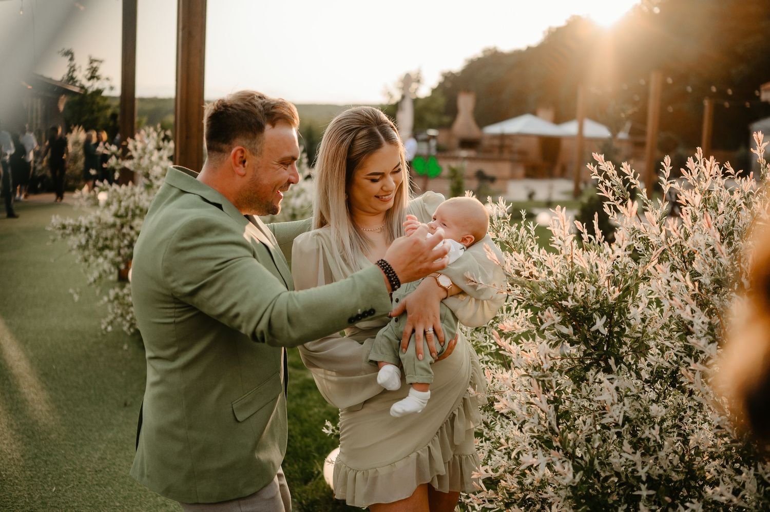 Smiling couple admires their newborn baby outdoors surrounded by white flowering bushes at sunset.