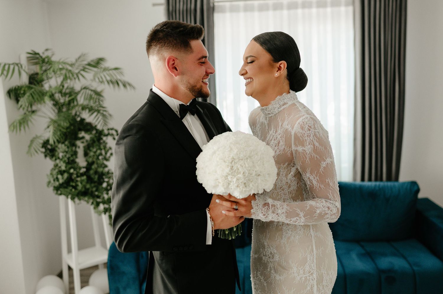 Bride and groom sharing a tender first look moment holding white floral bouquet indoors.