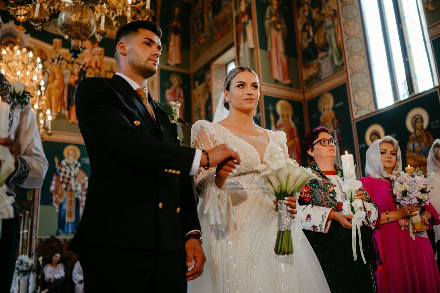Bride and groom standing at Orthodox church altar, bride holding calla lilies in elegant beaded wedding dress.
