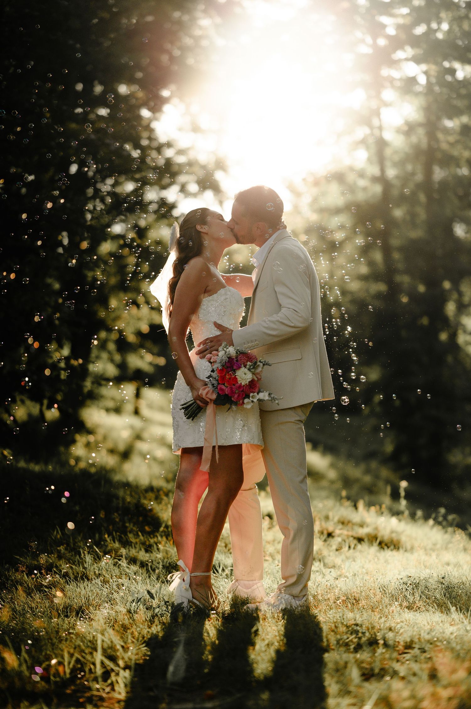 Couple sharing romantic kiss backlit by golden sunset light in forest during outdoor wedding celebration.