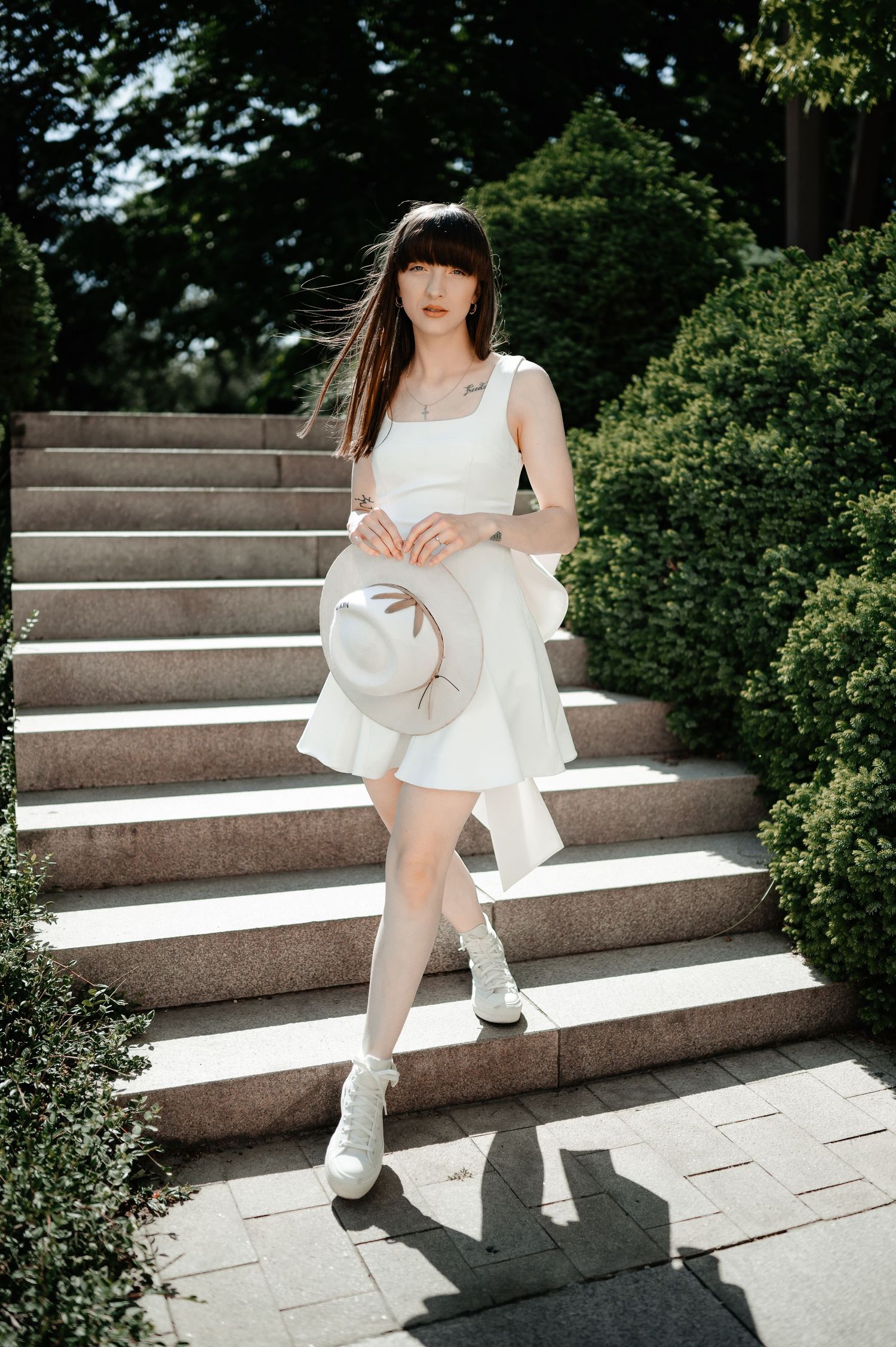 Elegant woman in white mini dress holding a hat while posing on sunlit stone steps outdoors.