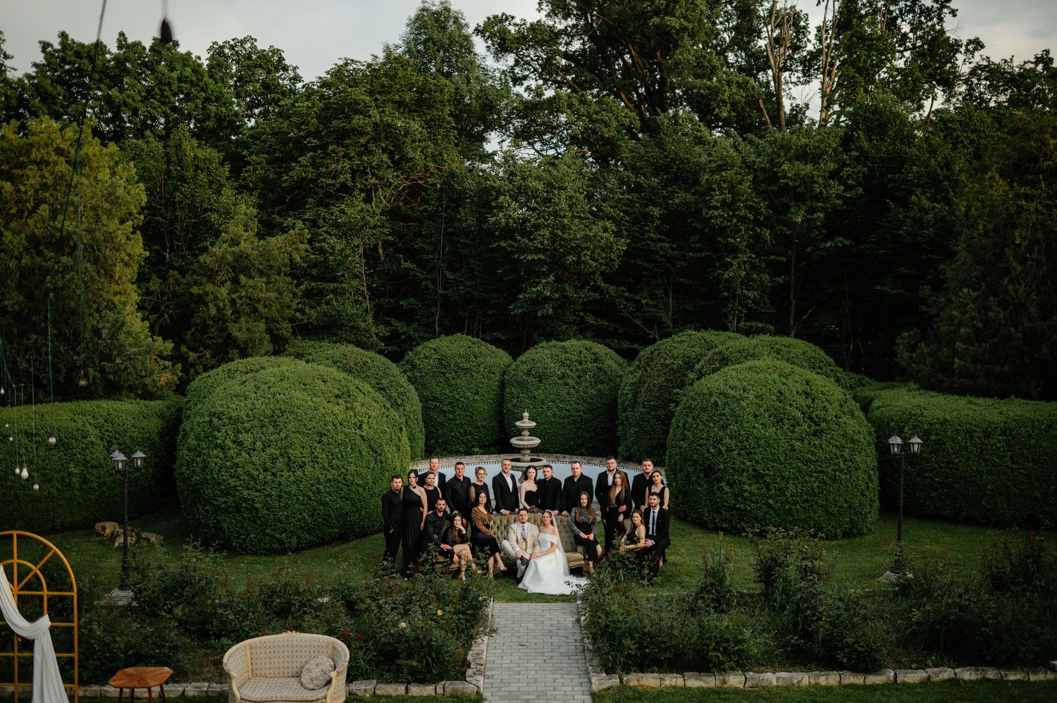Aerial view of wedding party gathered around a fountain surrounded by manicured topiary bushes.