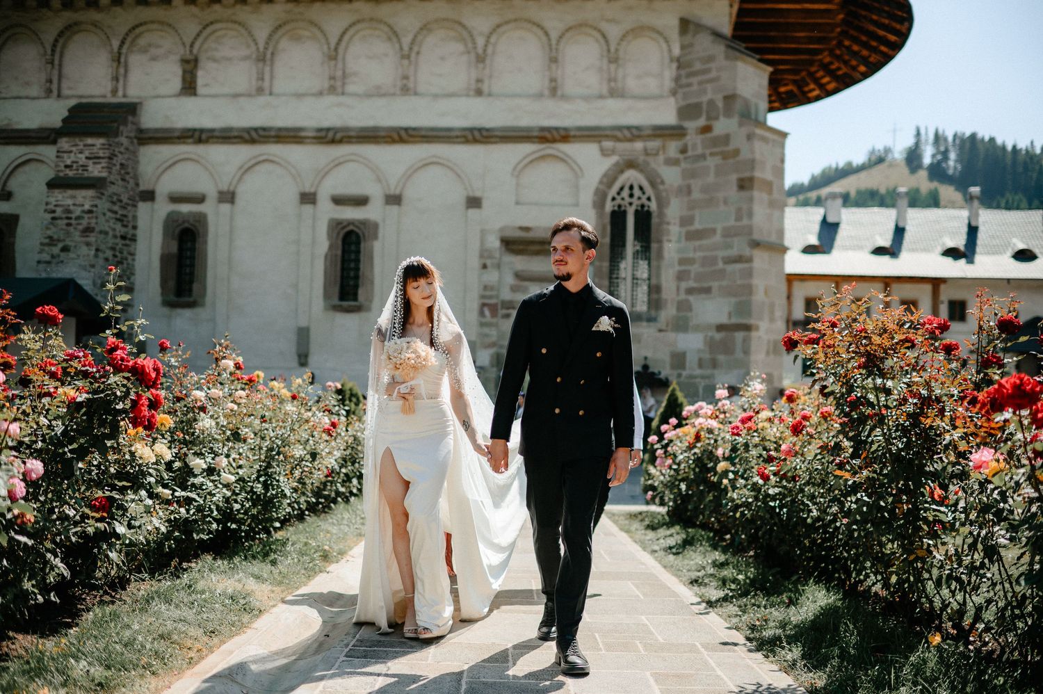 Bride and groom walk hand in hand through a rose garden at a historic monastery on their wedding day.