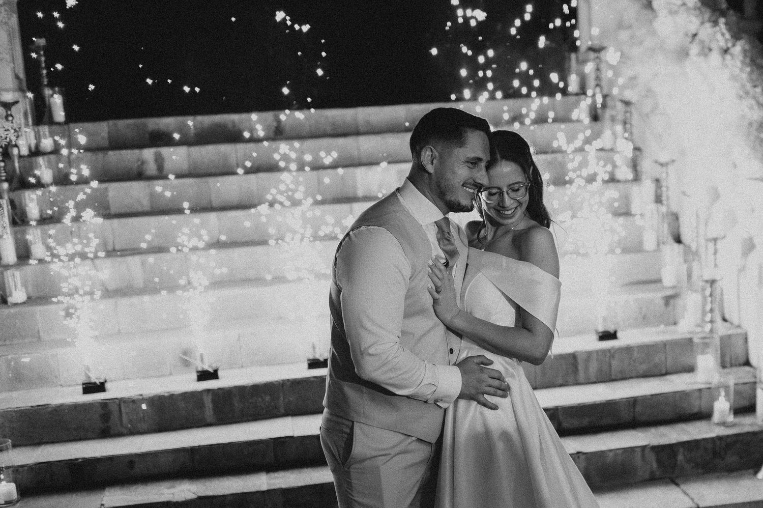 Newlyweds share tender embrace surrounded by spectacular fireworks display at elegant outdoor wedding reception.