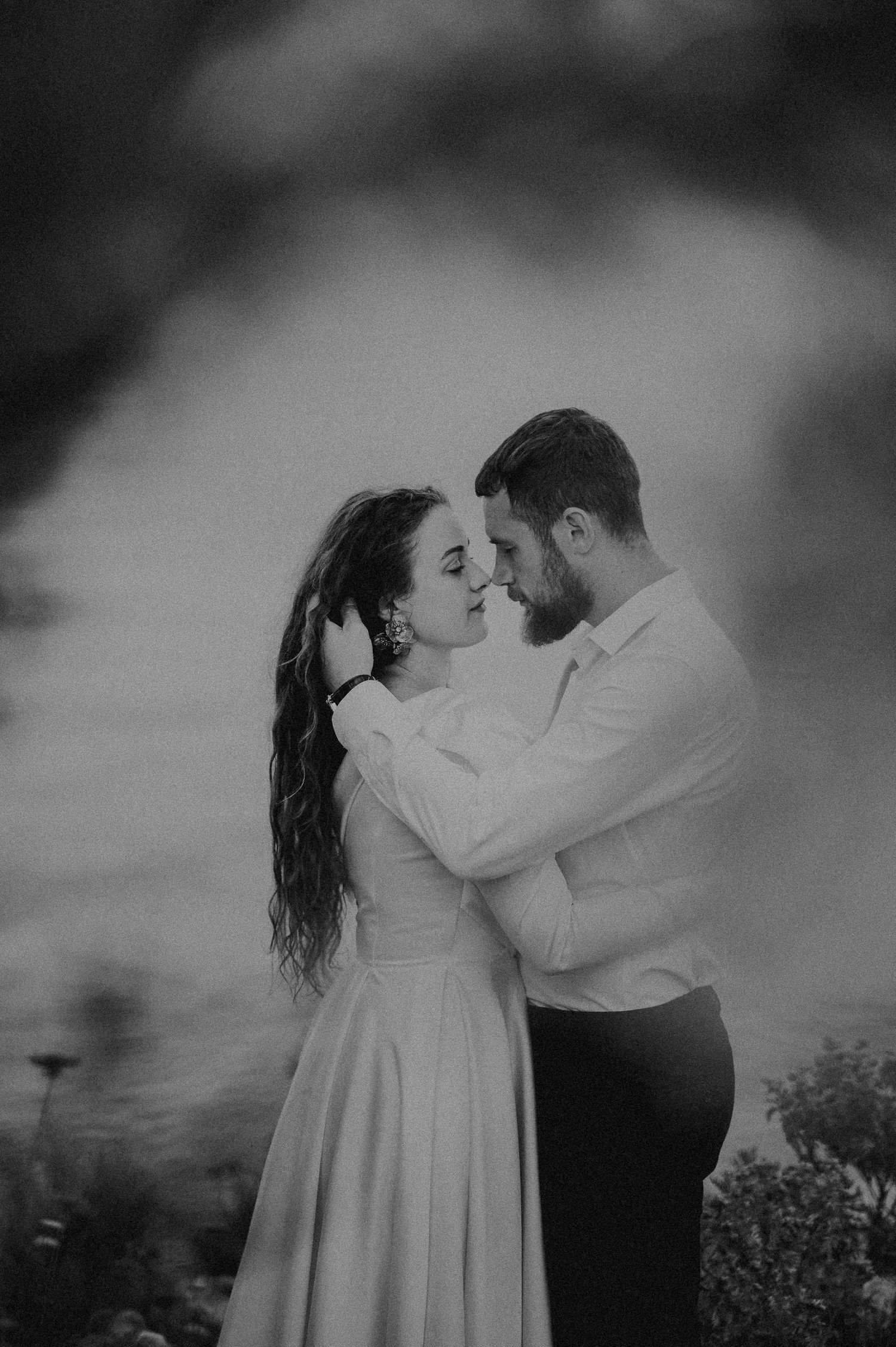Black and white intimate portrait of bride and groom nearly kissing while embracing outdoors at dusk.