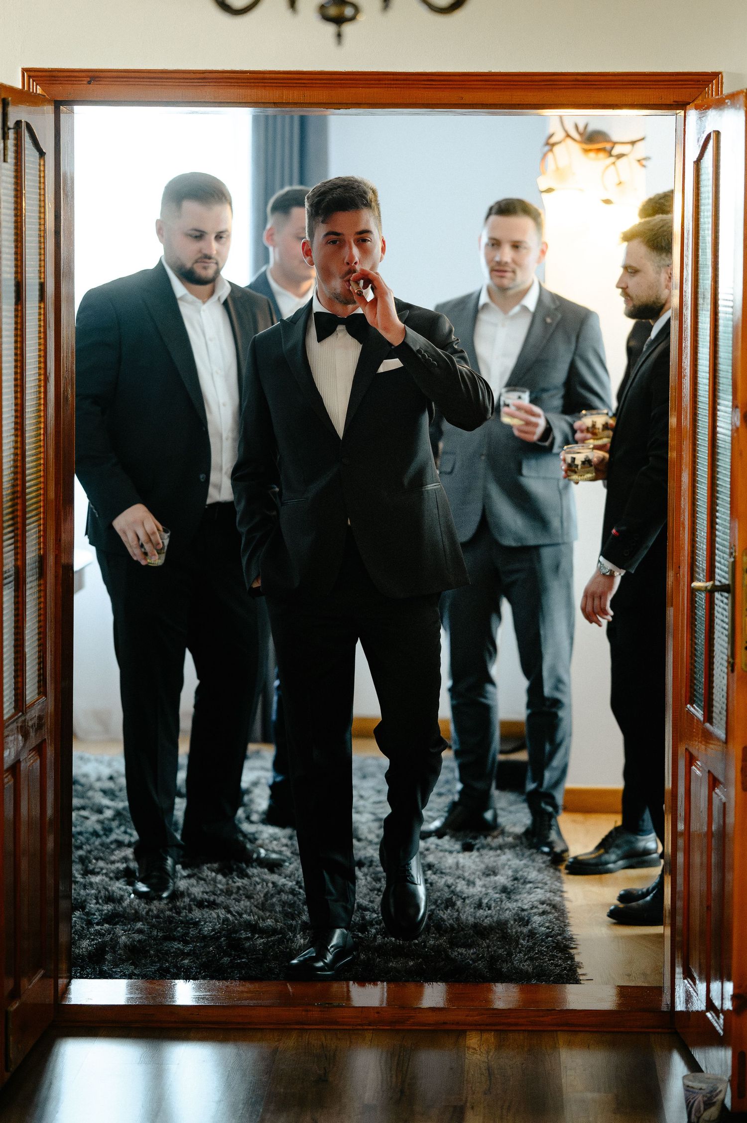Groom in black tuxedo walking through doorway with groomsmen behind him.