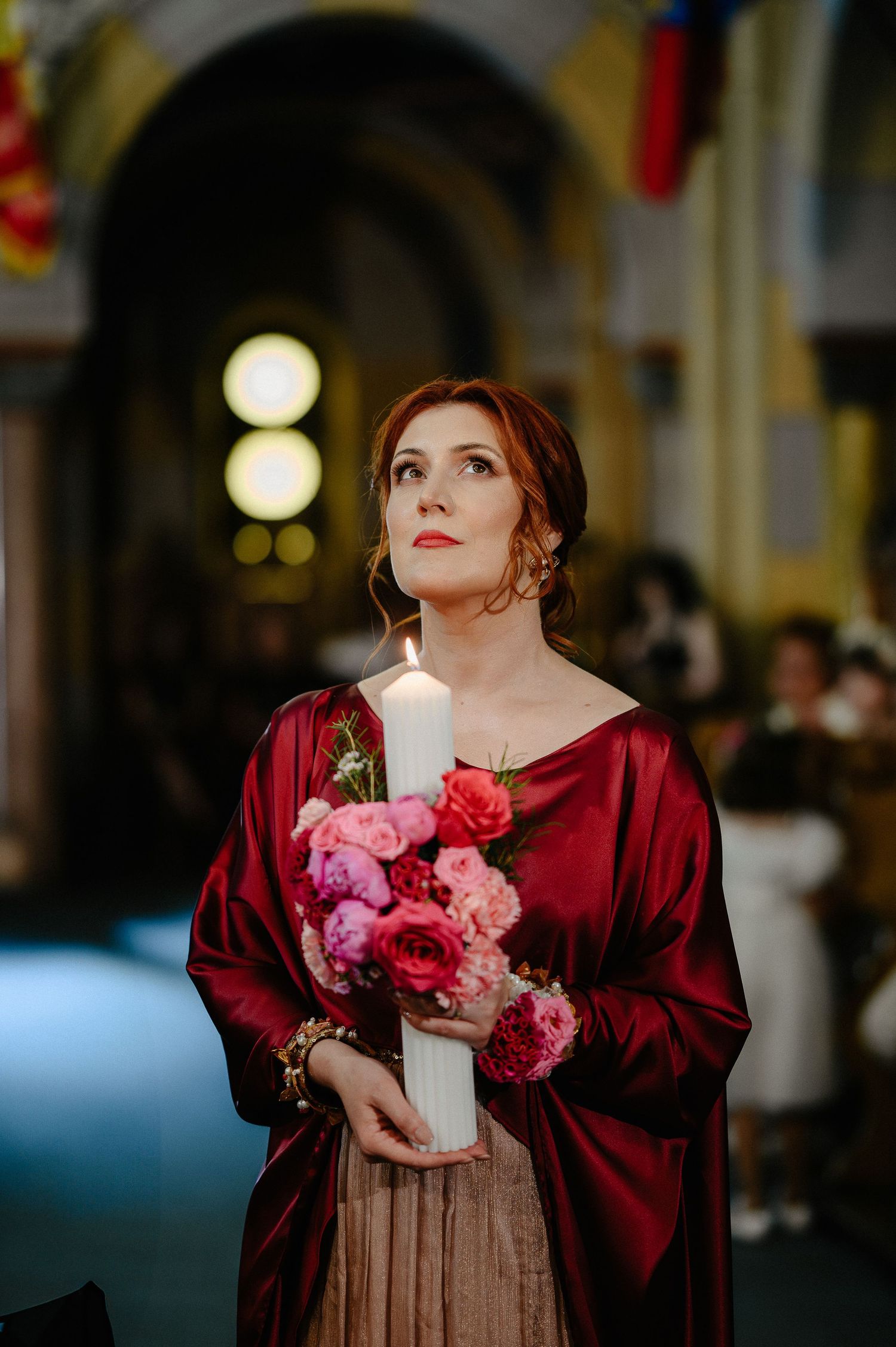 Elegant woman in burgundy wrap holding a large white candle and pink floral bouquet during a church ceremony.