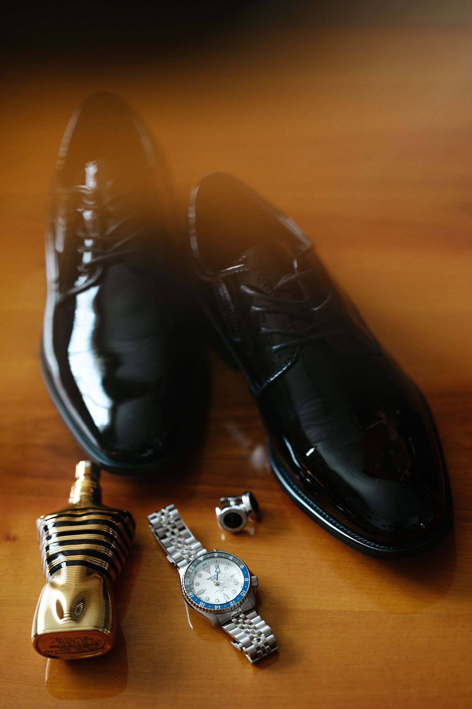 Shiny black patent leather dress shoes with a silver watch, cufflinks, and gold cologne bottle on wooden surface.