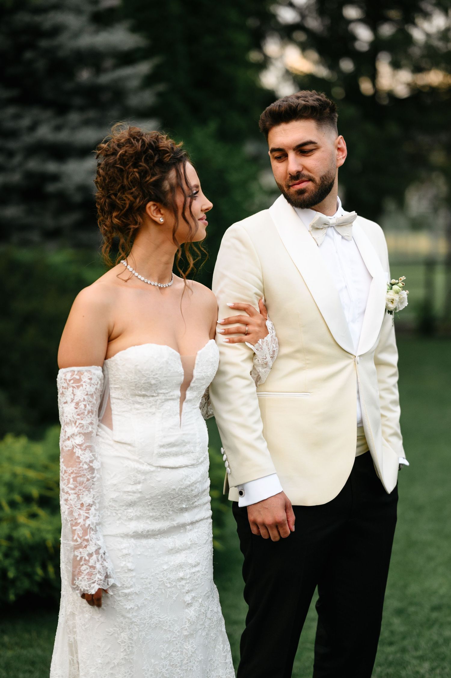 A bride in a white lace gown and groom in ivory tuxedo share a romantic moment outdoors on their wedding day.