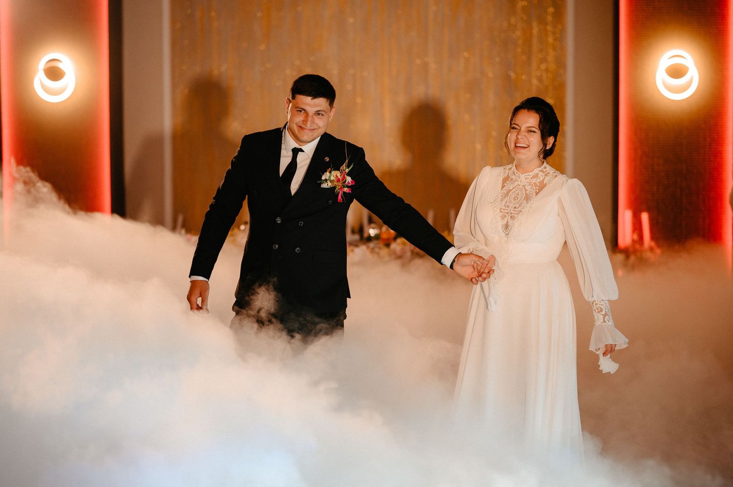 Newlyweds walk hand in hand through dramatic fog effect on wedding reception dance floor with red lighting.