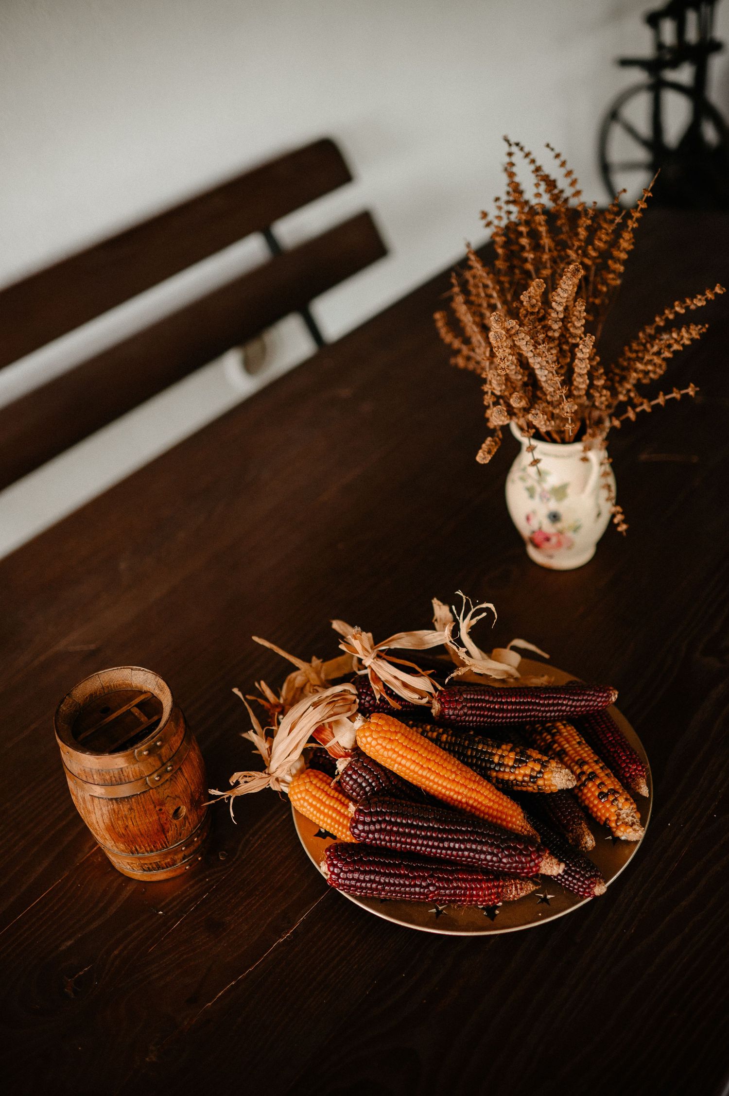 Autumn harvest display with colorful Indian corn on a plate, wooden mug, and dried wheat bouquet on a dark wood table.