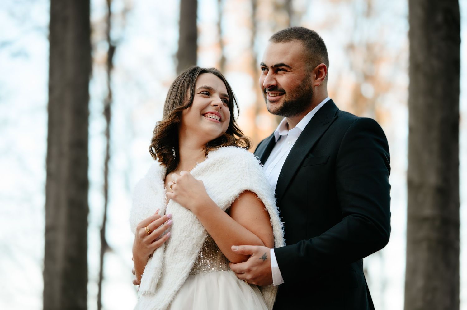Couple in wedding attire embracing outdoors among trees, bride in white fur wrap smiling at groom in black suit.