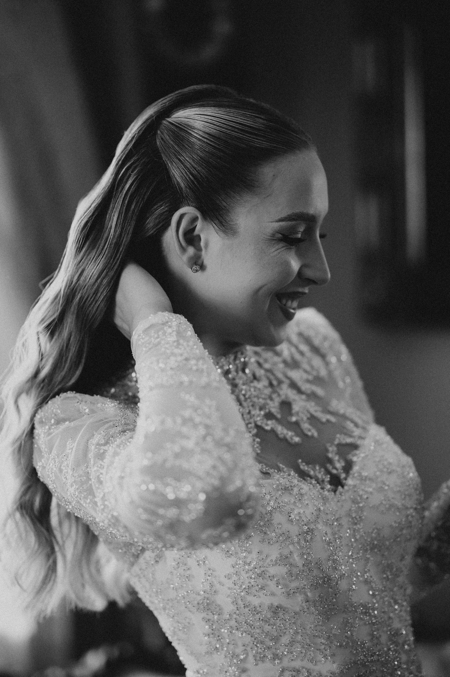 Black and white photo of a smiling bride in a beaded lace gown, adjusting her elegant half-up hairstyle.