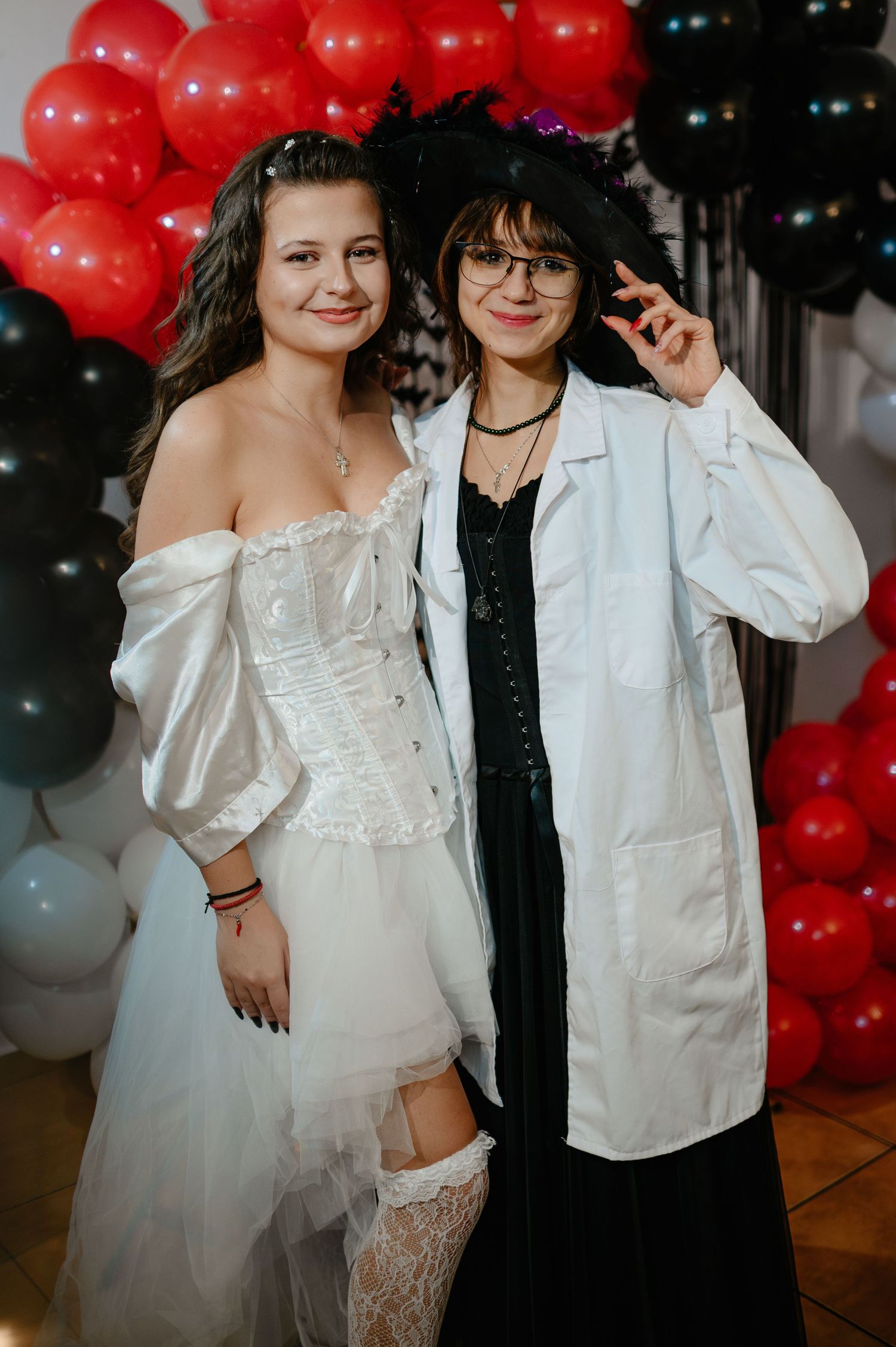 Two women in Halloween costumes smile together in front of red and black balloon decorations.